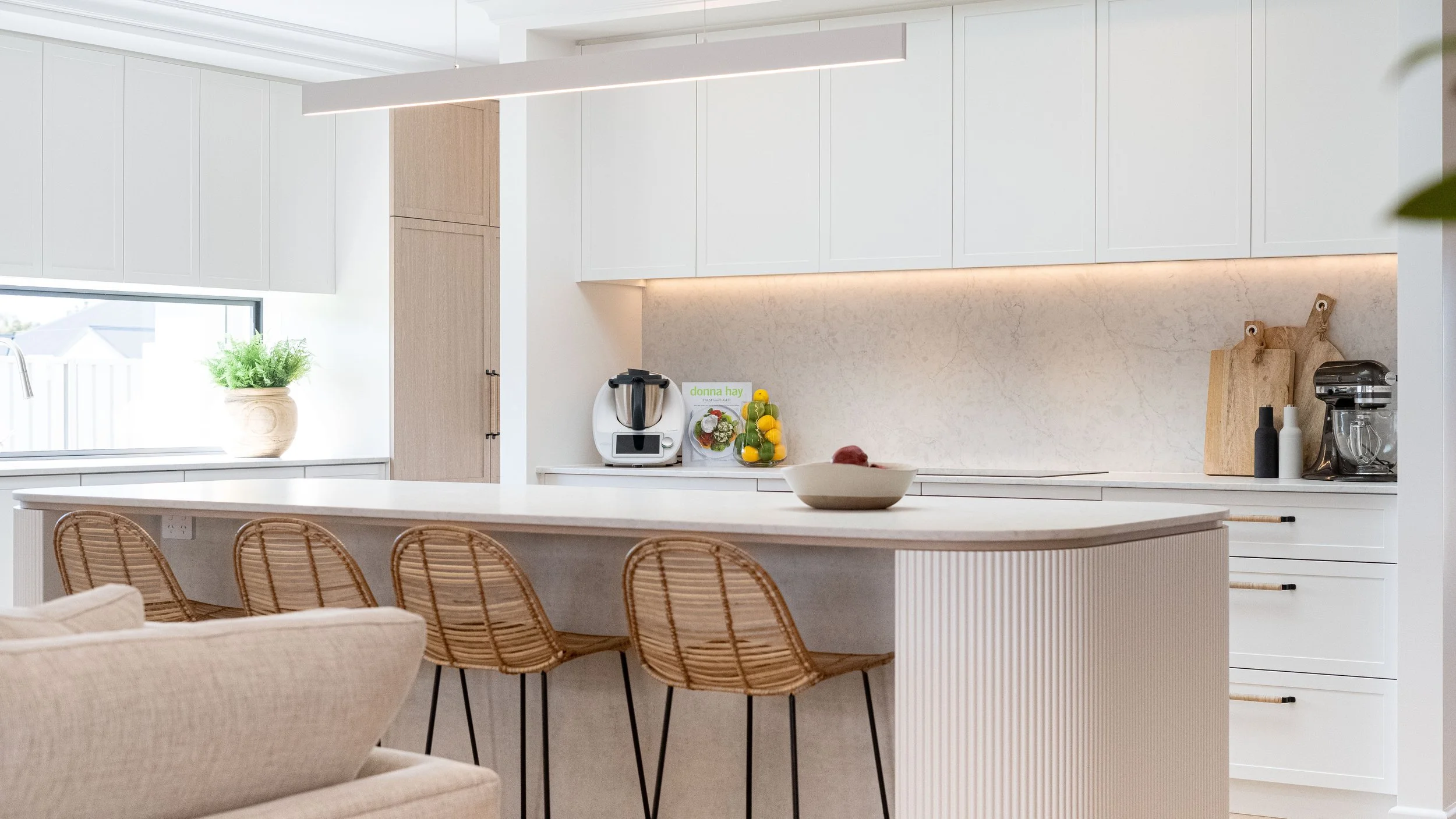 Modern kitchen with white cabinets, beige backsplash, and natural wood accents, featuring a kitchen island with woven chairs, a fruit bowl, and various small appliances.