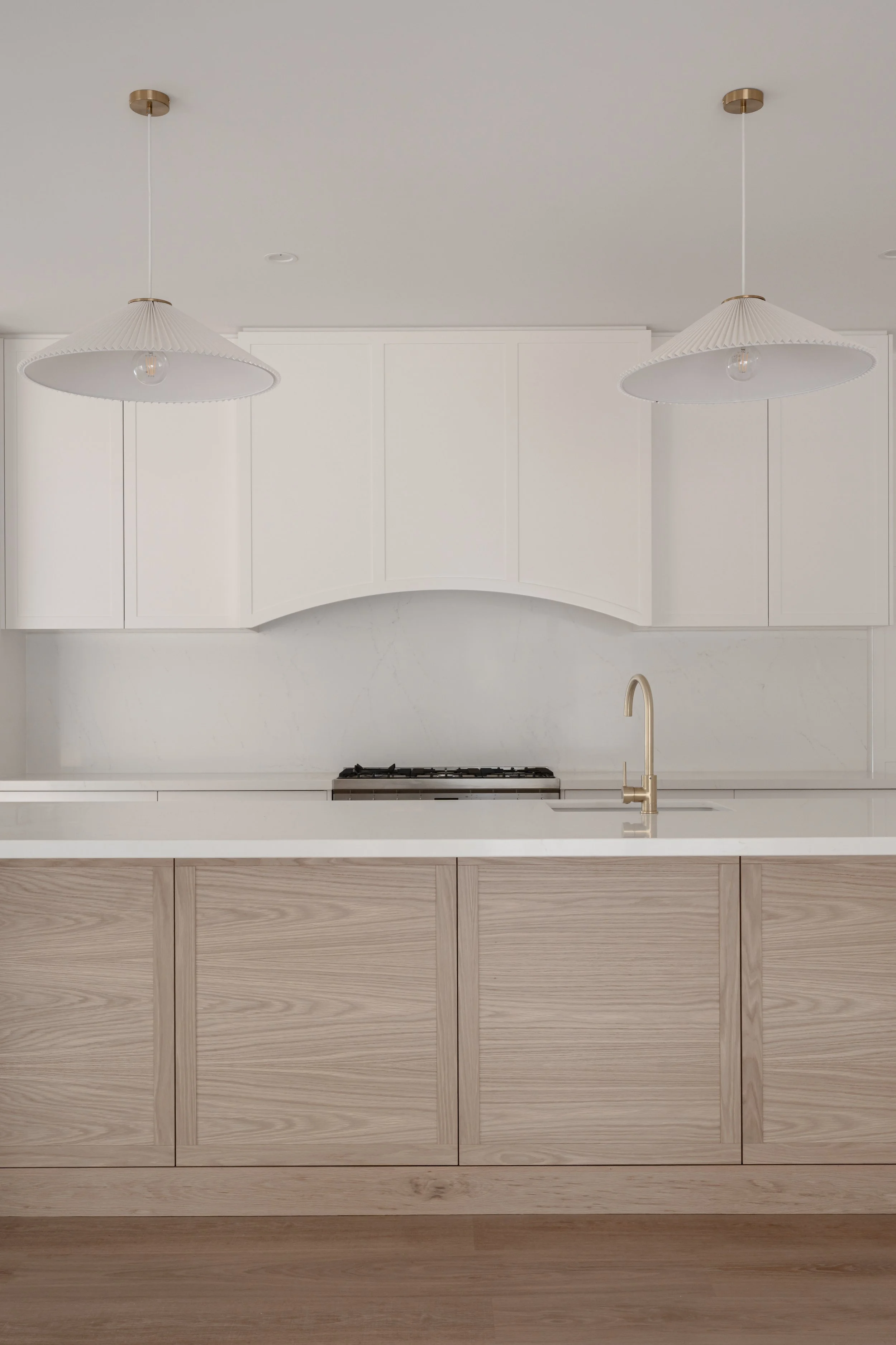Modern kitchen with white upper cabinets, a white countertop, and light wood lower cabinets. There is a gold faucet and a stove with a range hood. Two white pendant lights hang from the ceiling.