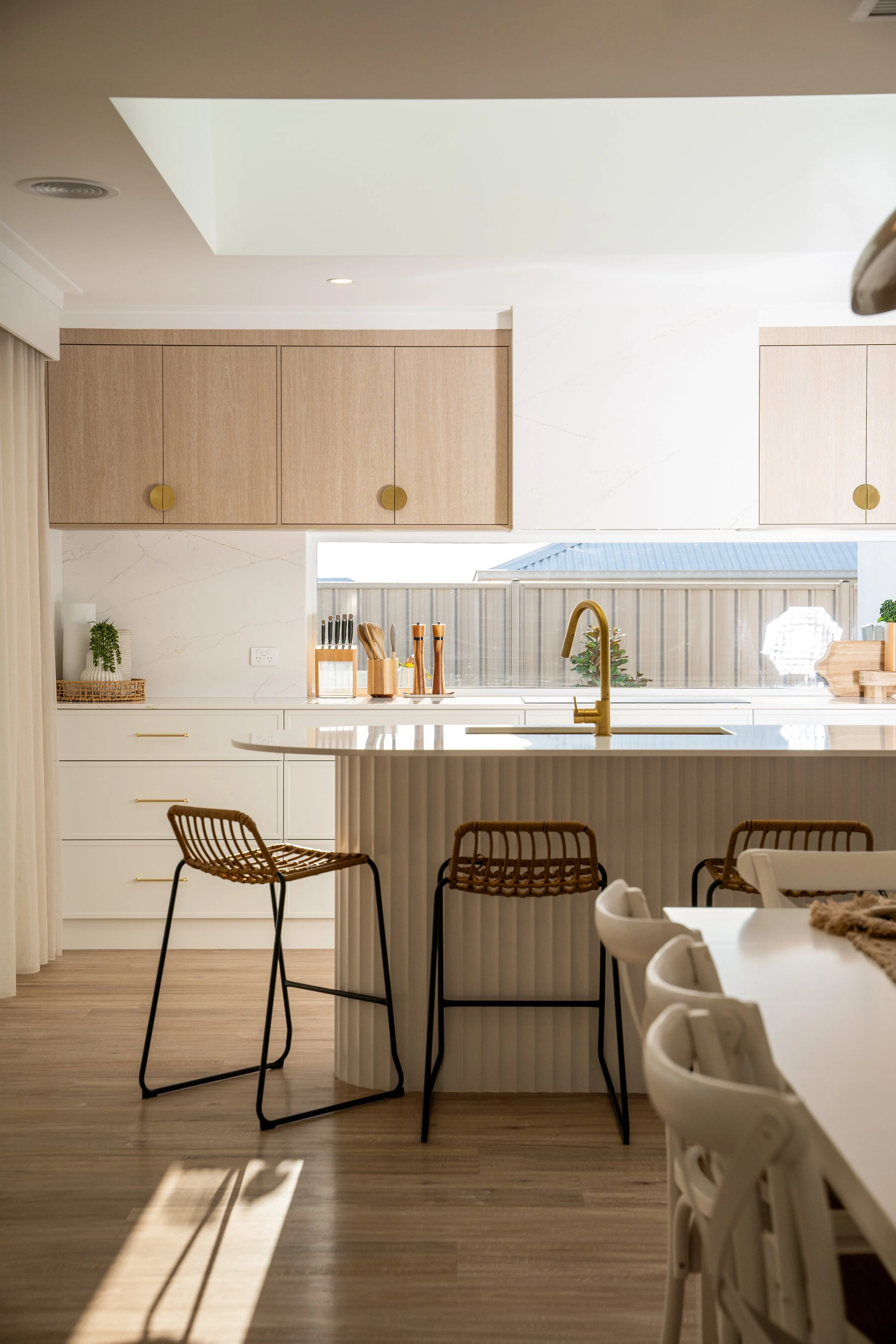 A modern kitchen with beige cabinets, white marble backsplash, and a large window. Kitchen island with bar stools, wooden utensils, and gold fixtures.