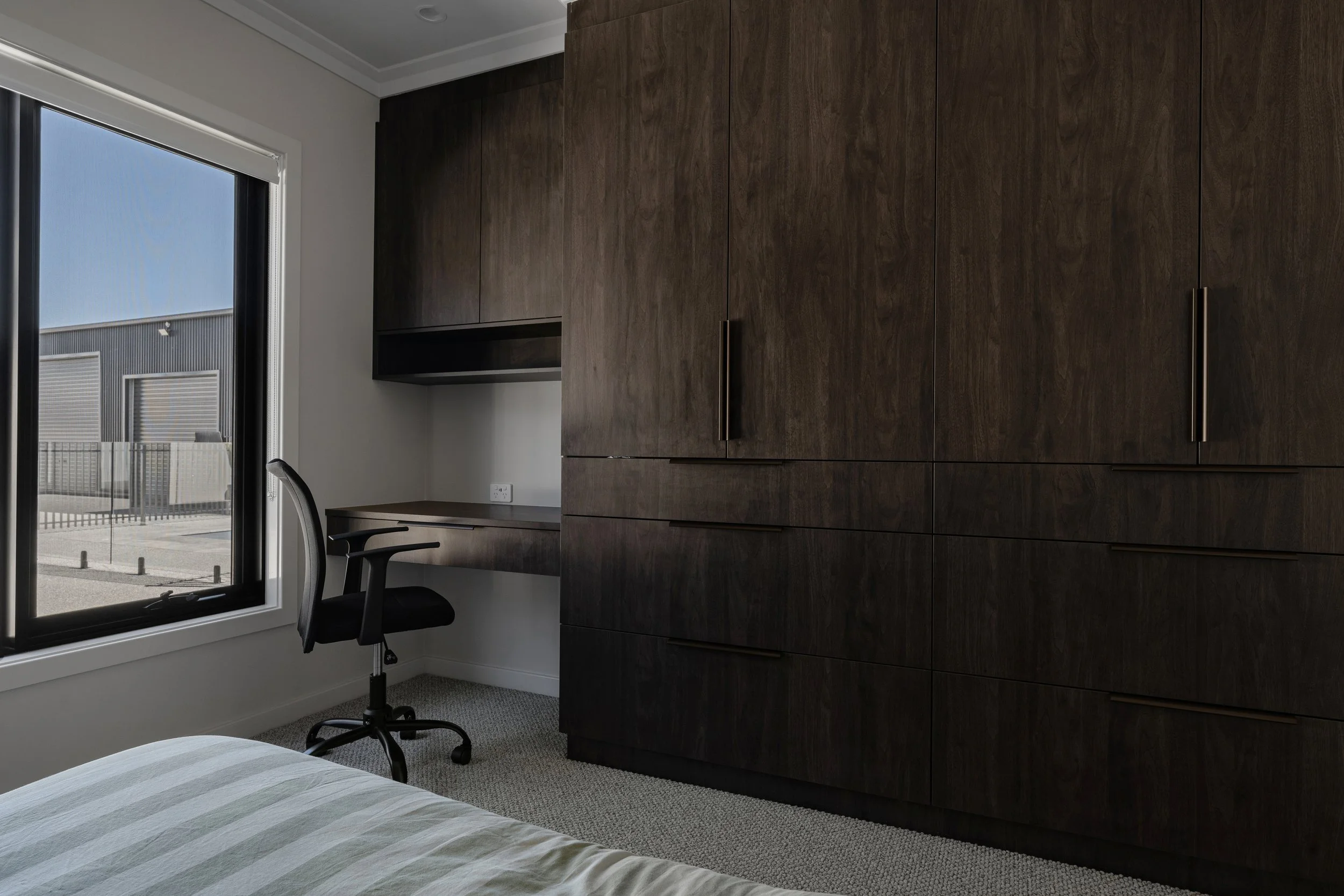 A modern bedroom with a large wooden wardrobe, a window with a view of an industrial building, a desk with a black office chair, and part of a bed with striped bedding.