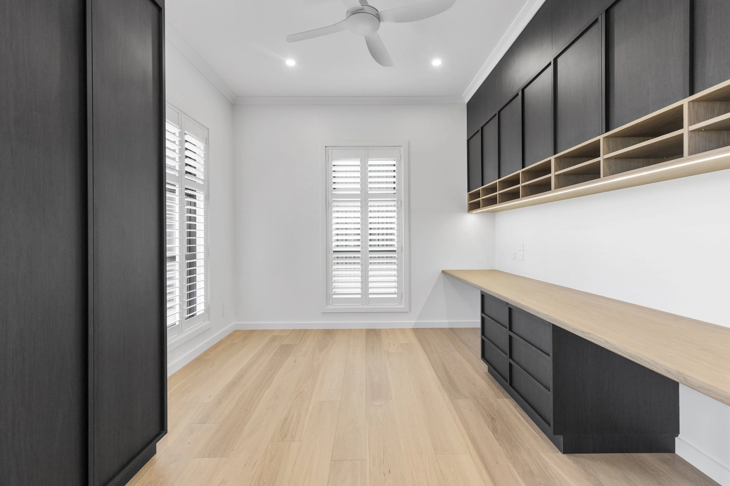 Empty room with black built-in cabinets, light wood floor, white walls, two windows with white shutters, ceiling fan, and recessed lighting.