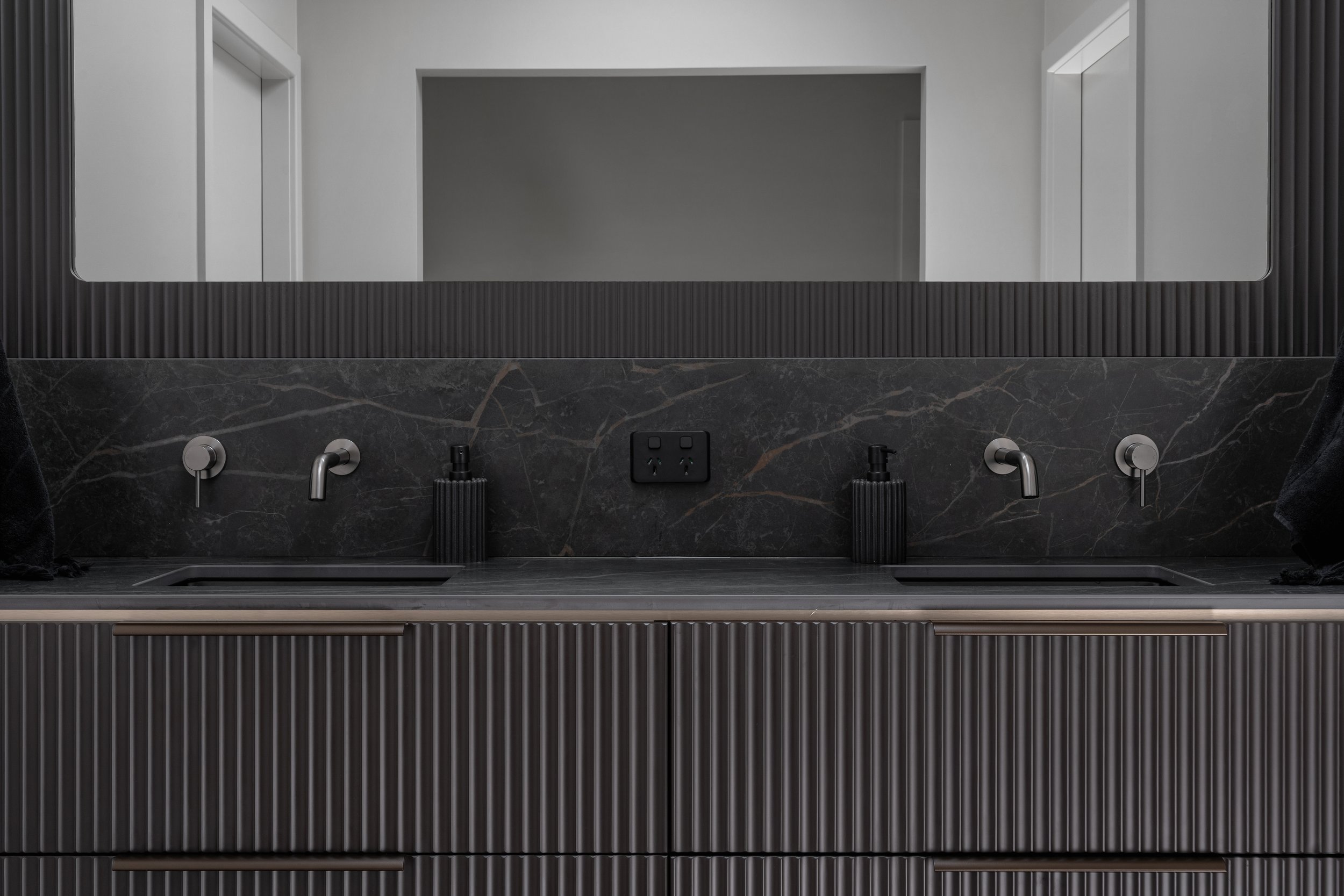 A modern bathroom vanity with a large rectangular mirror, two faucets, and black hand soap dispensers on a black marble countertop with gray veining. The wall and mirror frame are gray, and the vanity has a textured black finish.