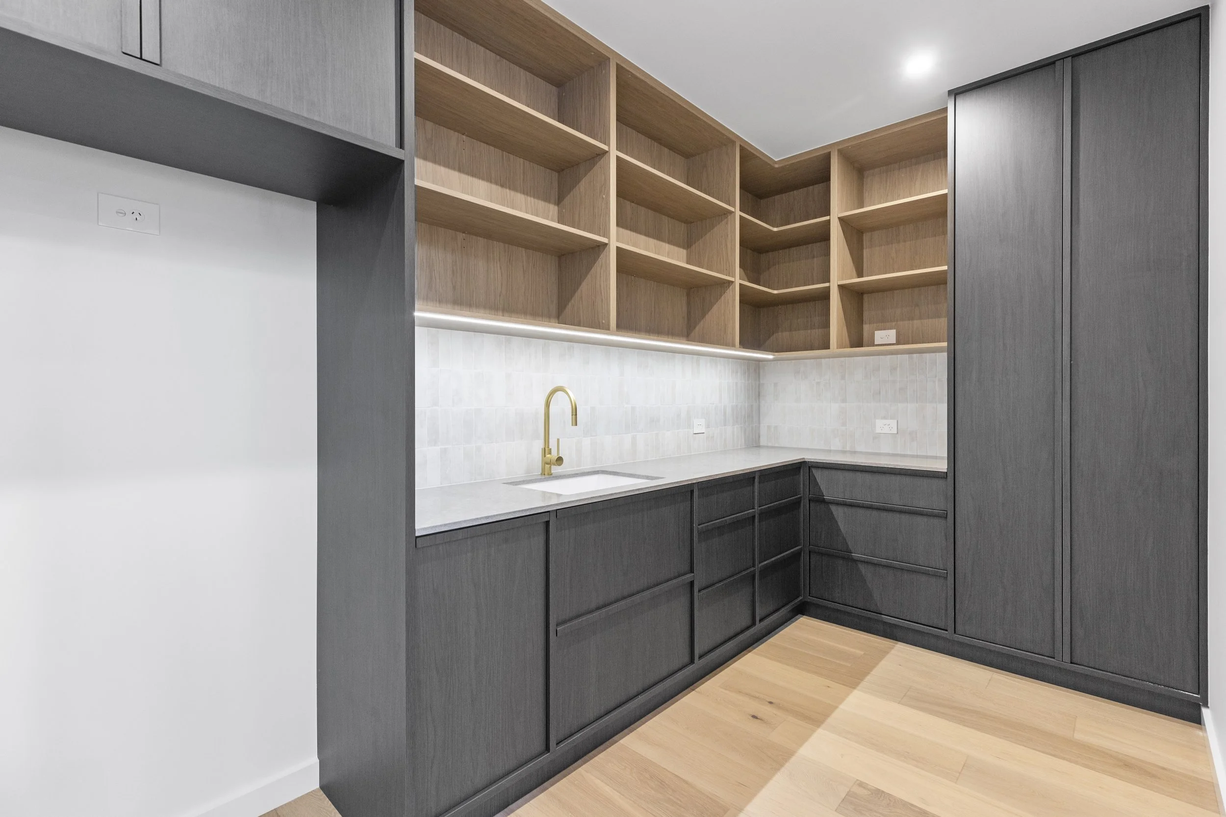 Modern kitchen with black cabinetry, wooden open shelves, a white countertop, a gold faucet, and wood flooring.