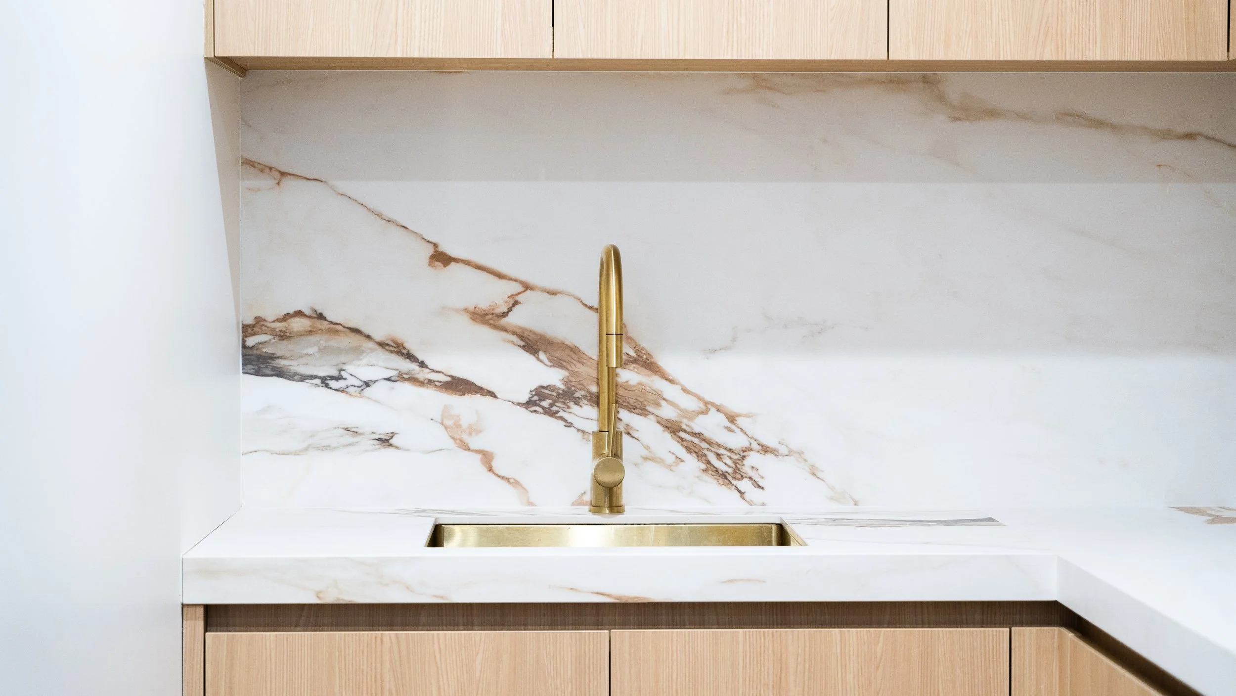 A modern kitchen sink area with a white marble backsplash featuring brown and black veining, a gold faucet, and a white countertop with a built-in sink.