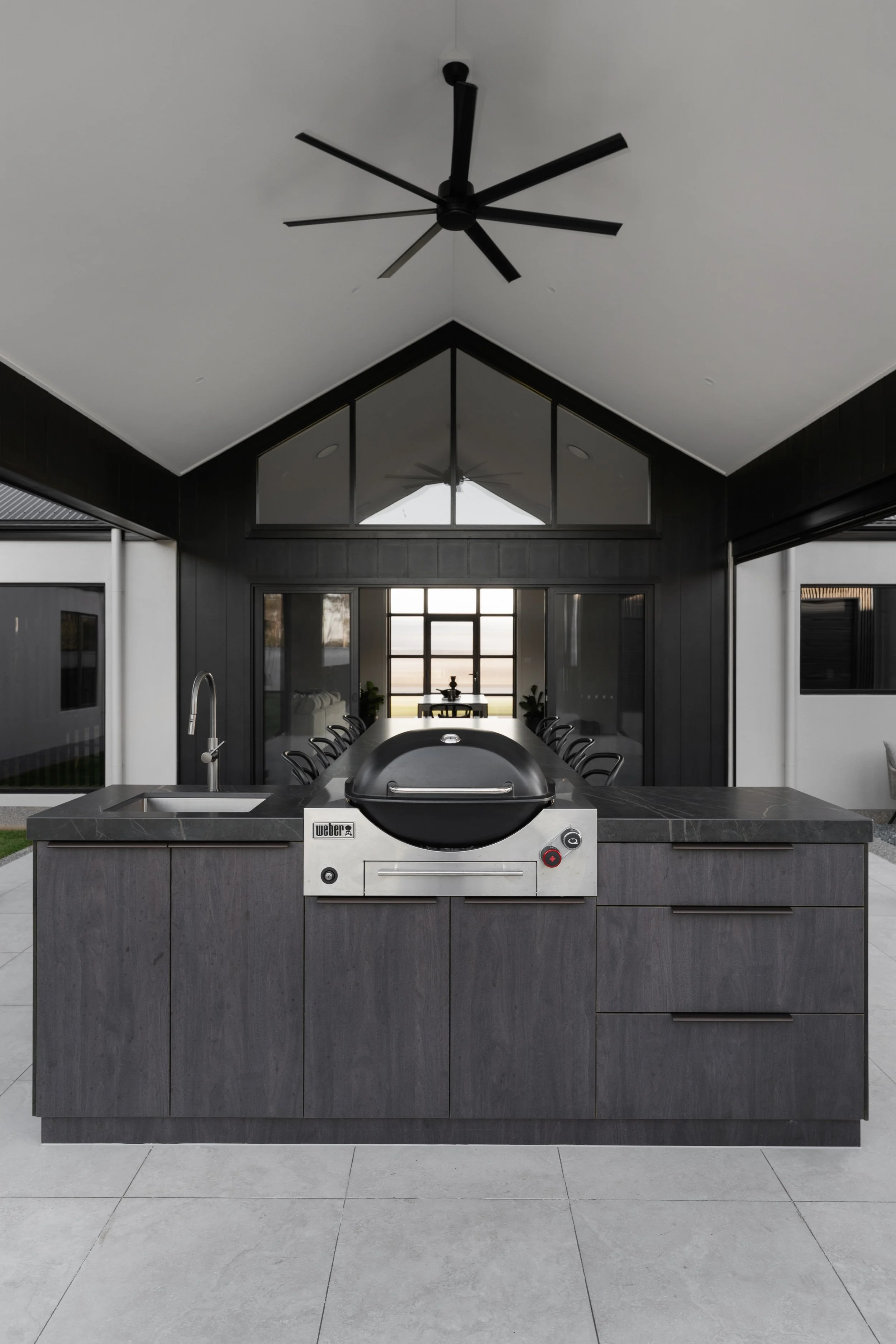 Modern outdoor kitchen with a large island featuring a built-in Weber grill and a black countertop, set against a backdrop of large windows and a view of the outdoors.