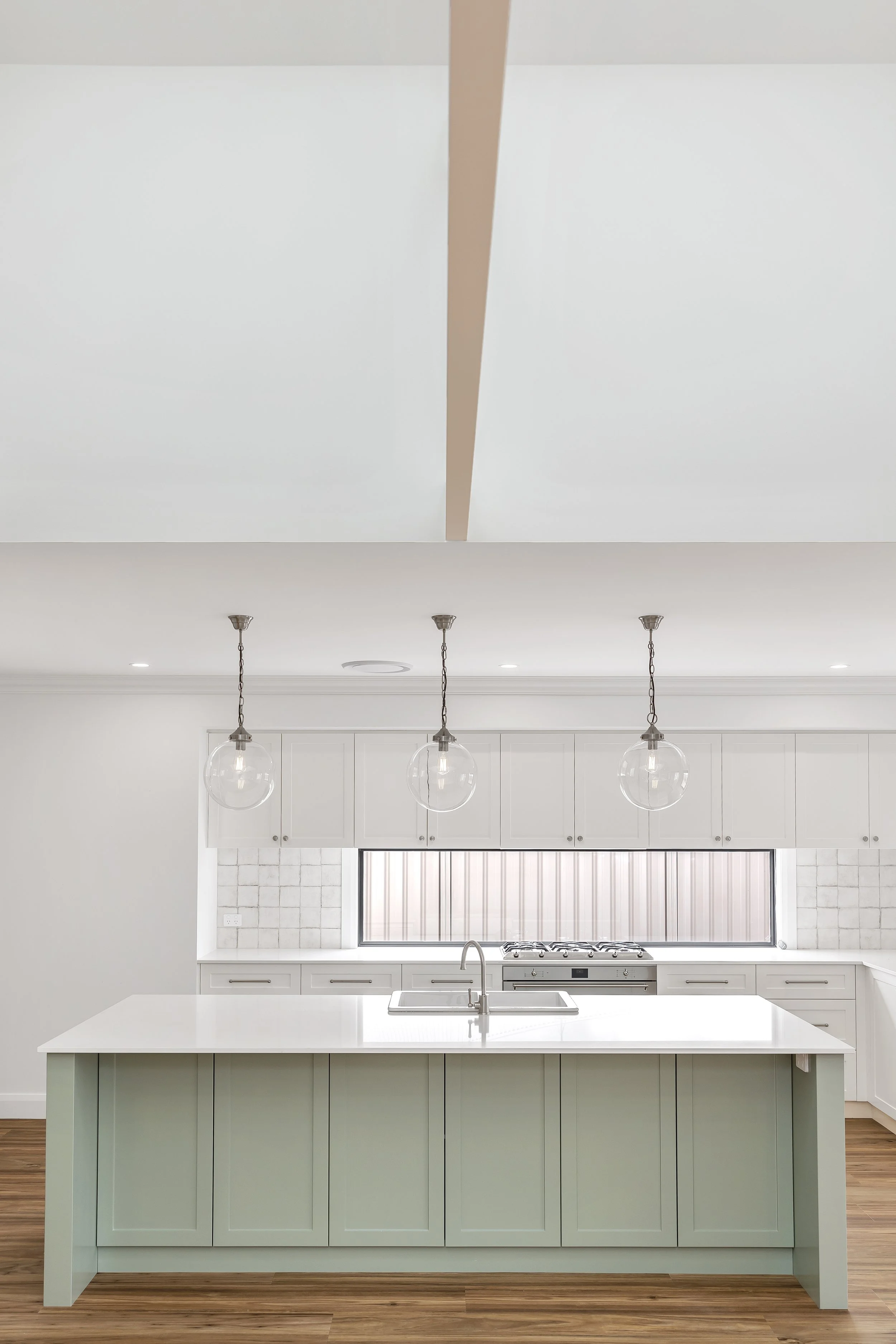 A modern kitchen with white cabinetry, a large kitchen island with a white countertop, a sink, hanging pendant lights, and a window with a gray blind. The kitchen has a hardwood floor and a clean, minimal decor style.