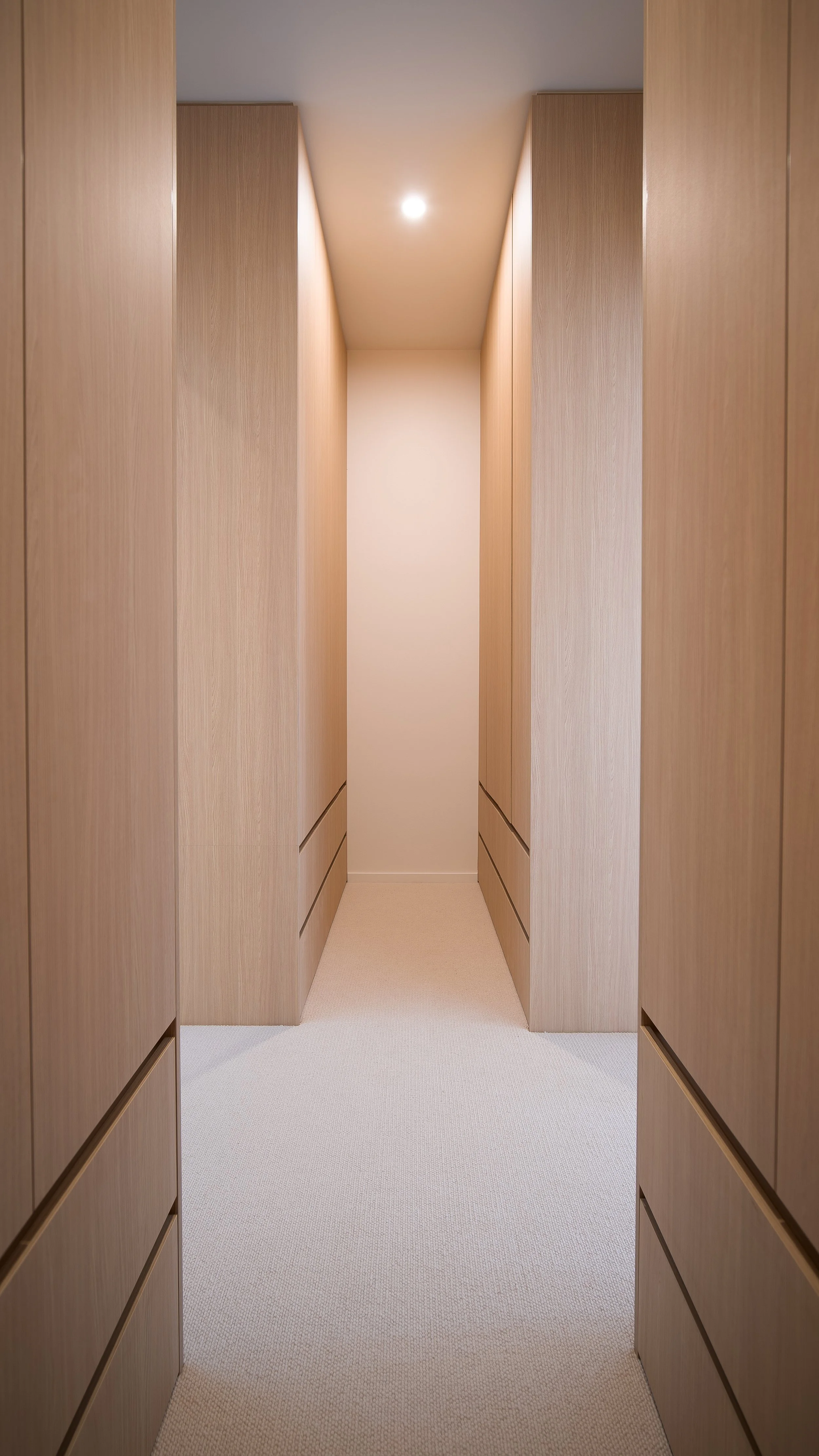 A narrow corridor with wooden paneling on both sides and a light-colored wall and ceiling. There is a small, round ceiling light in the center of the ceiling.