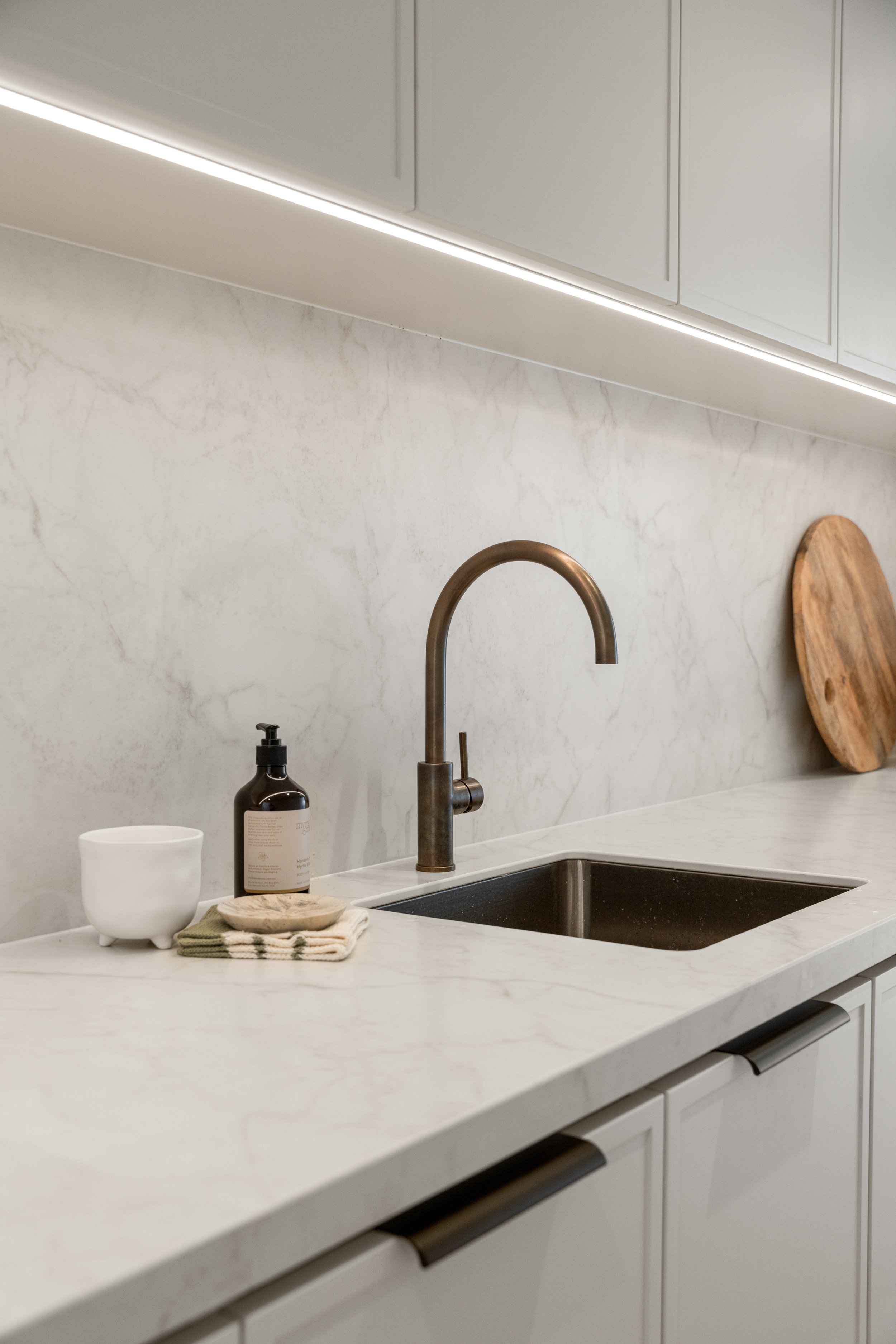 A modern kitchen countertop with a stainless steel sink, a black soap dispenser, a white mug, a small wooden plate, and neatly folded cloths. There are white cabinets above with sleek handles, and a wooden cutting board leaning against the marble bac