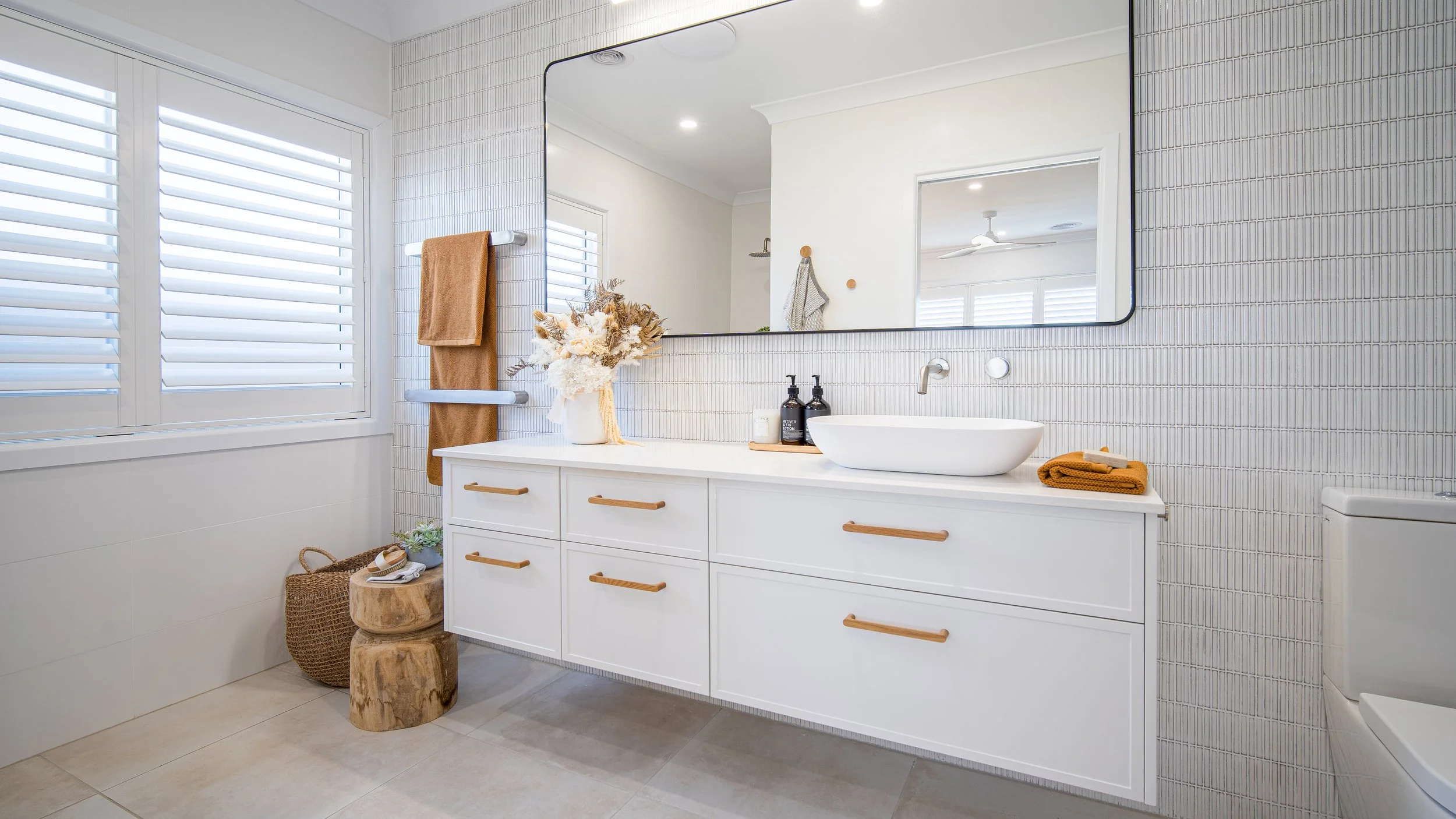 Modern bathroom with white vanity, large mirror, decorative dried flowers, folded towels, soap dispenser, and bidet, with natural light from windows with white shutters.