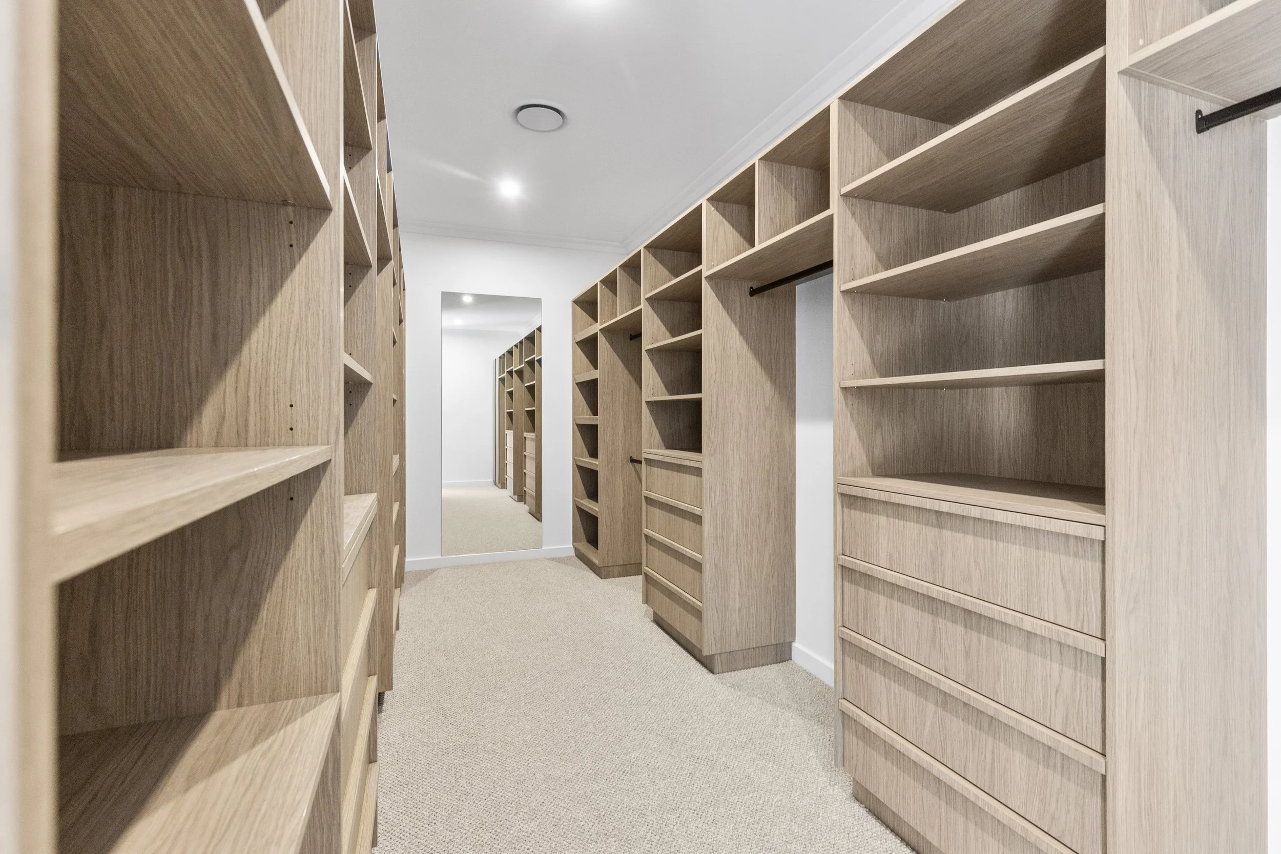 Empty wooden bookshelves in a walk-in closet with a mirror at the end and a ceiling light.