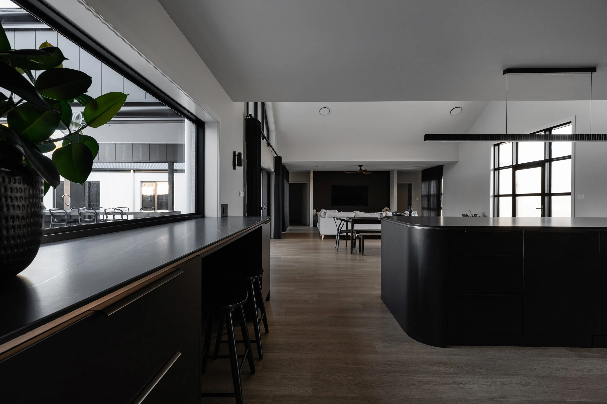 Interior view of a modern, minimalist living space with large windows, a gray kitchen island, black accents, and a glimpse of a dining area and living room with a TV and white sofa.