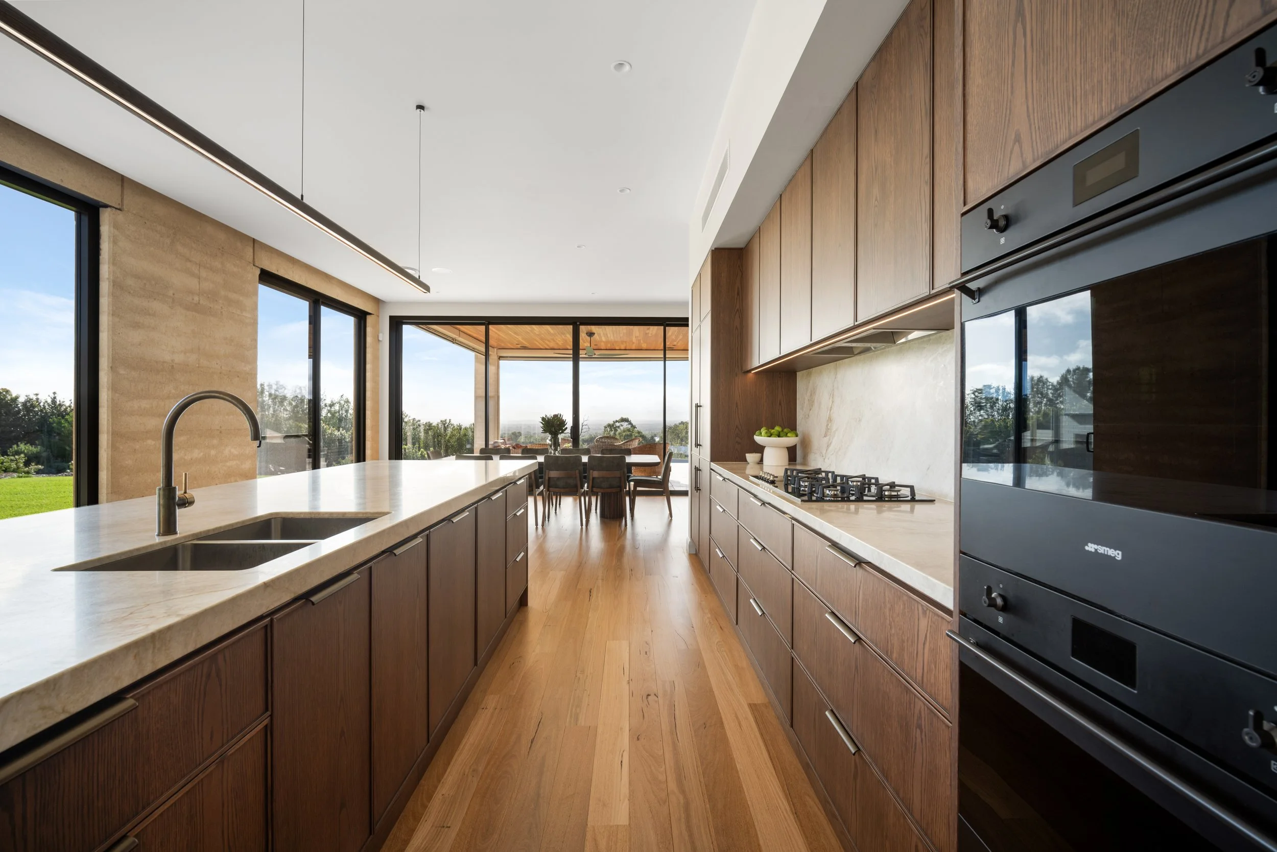Modern kitchen with wooden cabinets, marble countertops, large windows, and a view of a backyard with grass and trees.