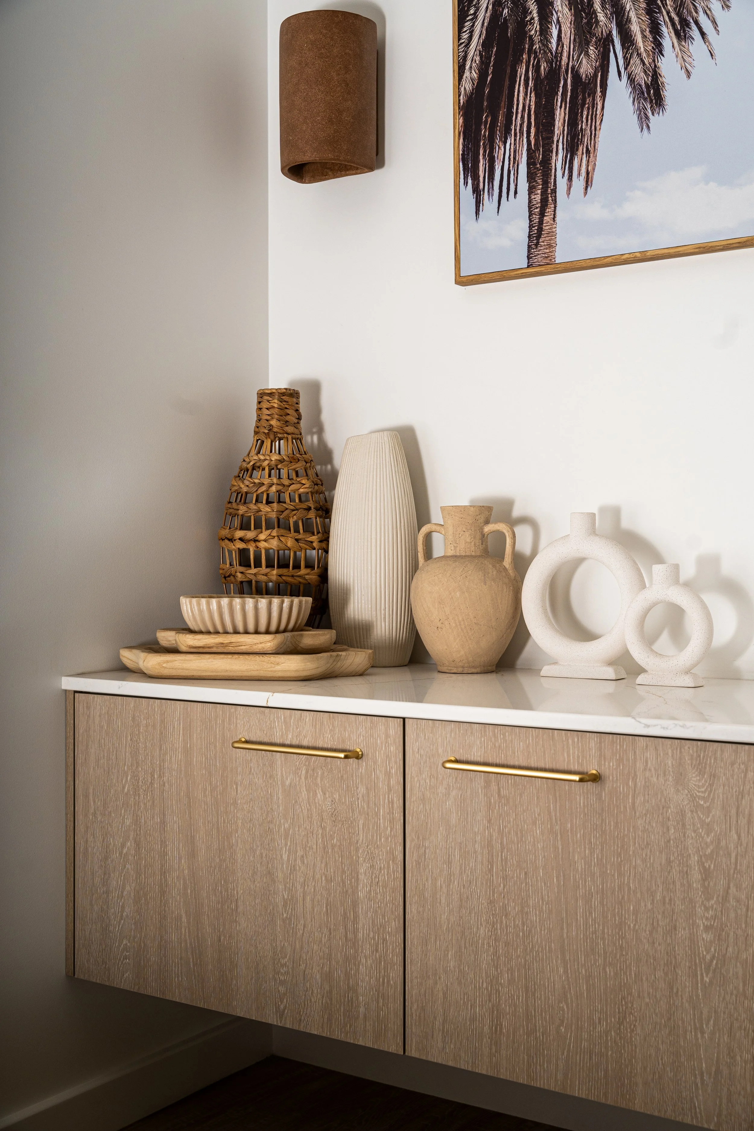 A light-colored wooden cabinet with two doors and gold handles, topped with decorative vases, bowls, and a woven basket, with a white wall in the background and framed art featuring a palm tree.