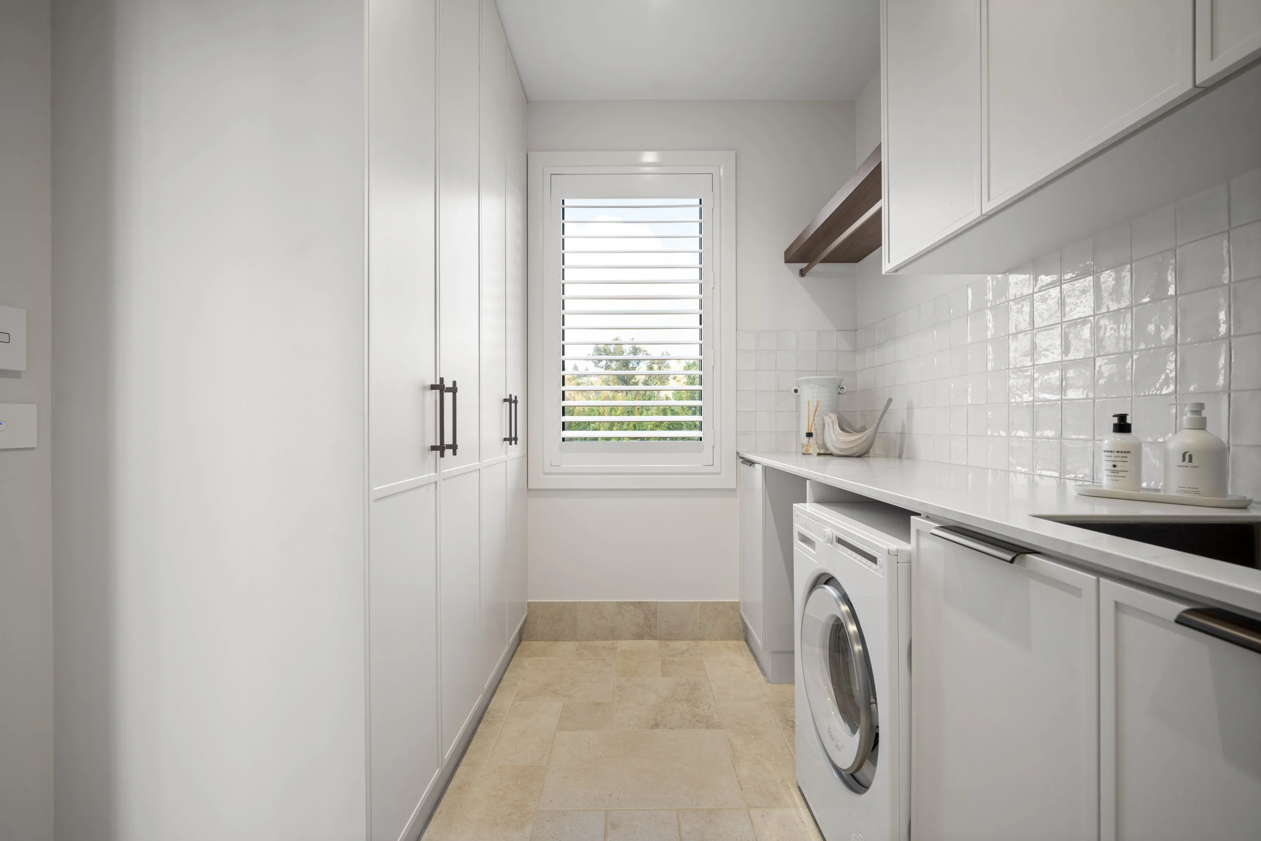 A small, white laundry room with a window covered by white horizontal blinds. There are white cabinets on one side, a countertop with soap bottles, and a washing machine underneath. The floor is tiled with beige tiles.