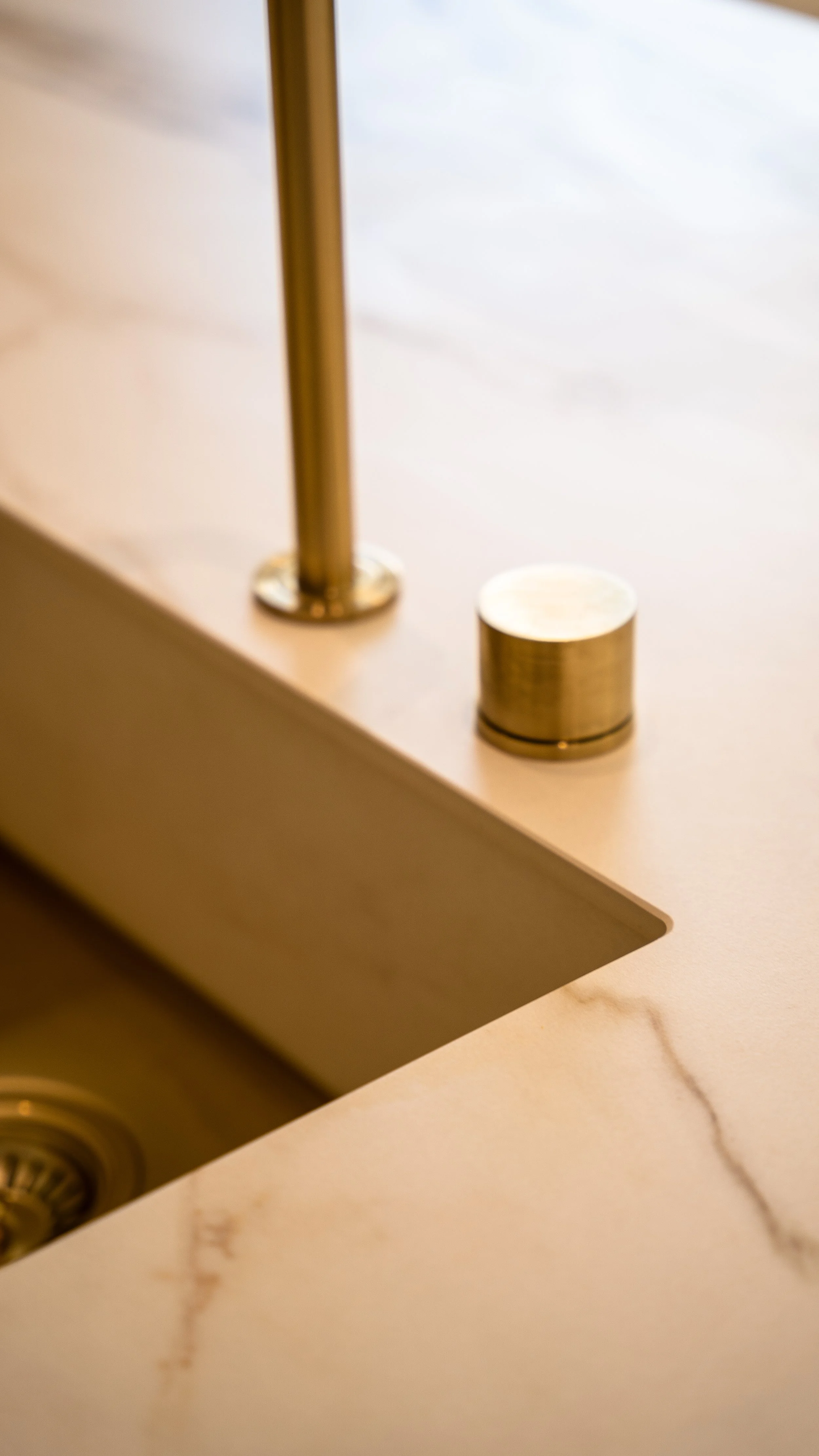Close-up of a gold-colored faucet and drain in a kitchen sink with a beige marble countertop.