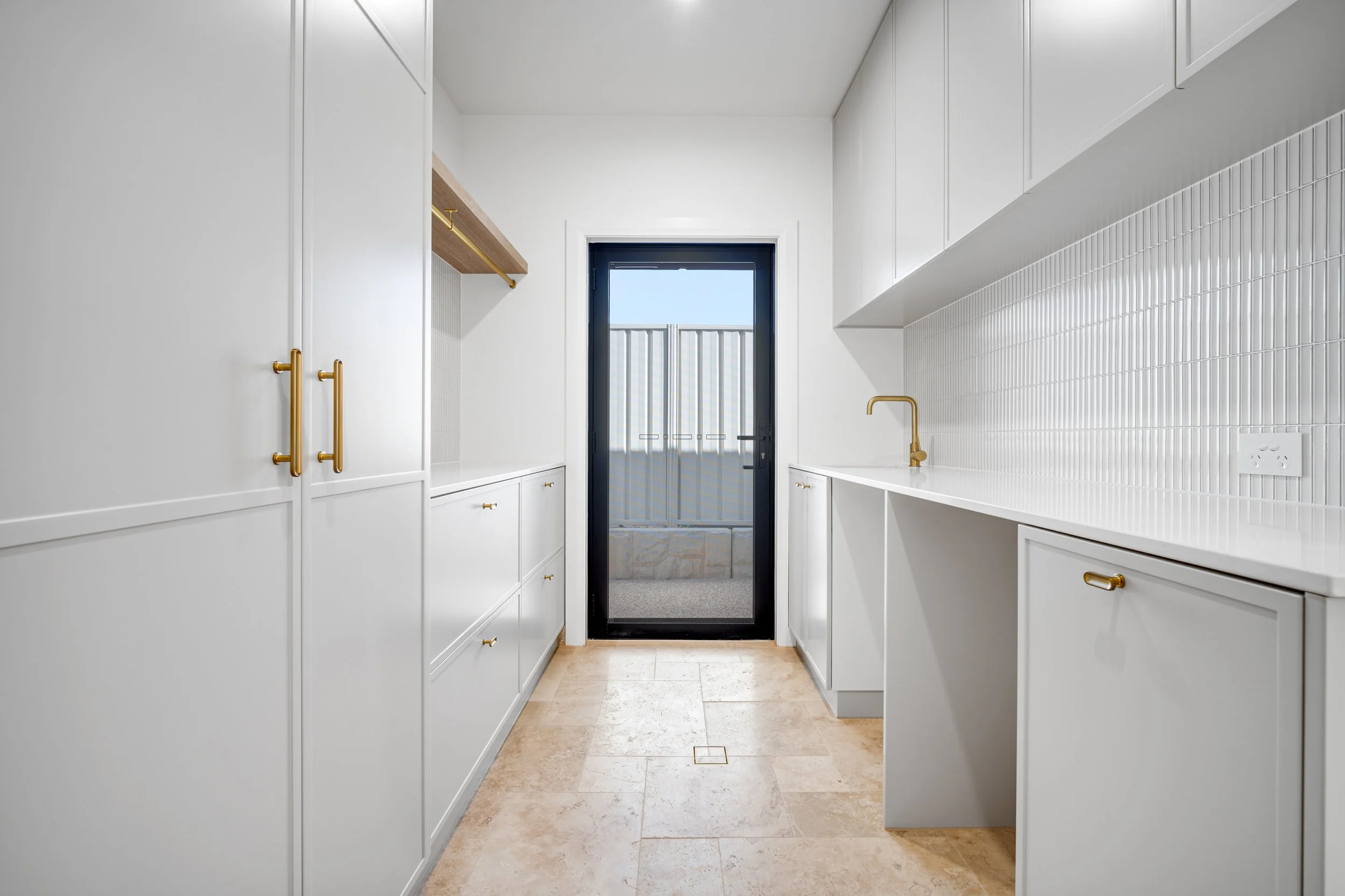 Modern white kitchen with gold hardware, a small wooden shelf above, and a black door leading to an outdoor patio with a metal railing.