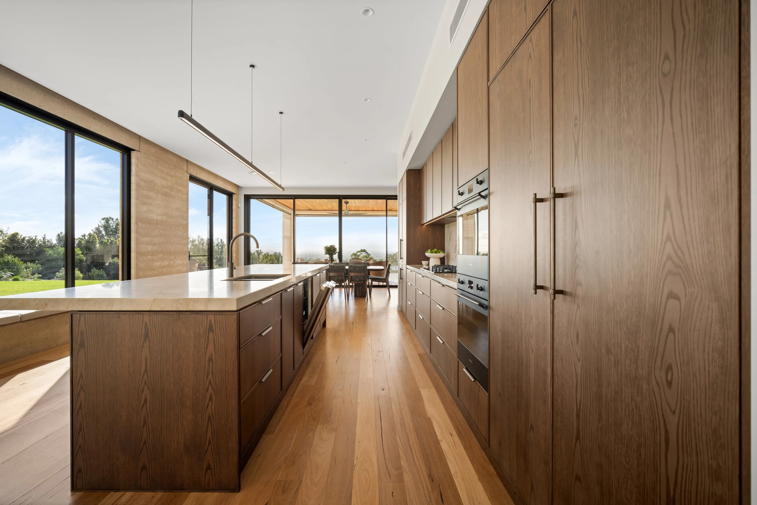 Modern kitchen interior with wooden cabinets, marble island, stainless steel appliances, and large windows showing a green outdoor landscape.