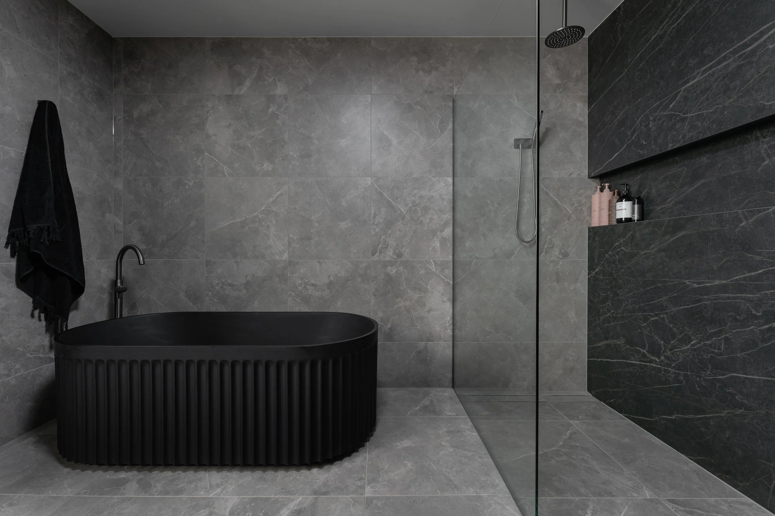 Modern bathroom with grey marble tiles, a black freestanding bathtub, a black wall-mounted faucet, a black towel hanging on a hook, a large mirror, a walk-in shower with a rainfall showerhead, and a built-in shelf with shampoo and soap bottles.