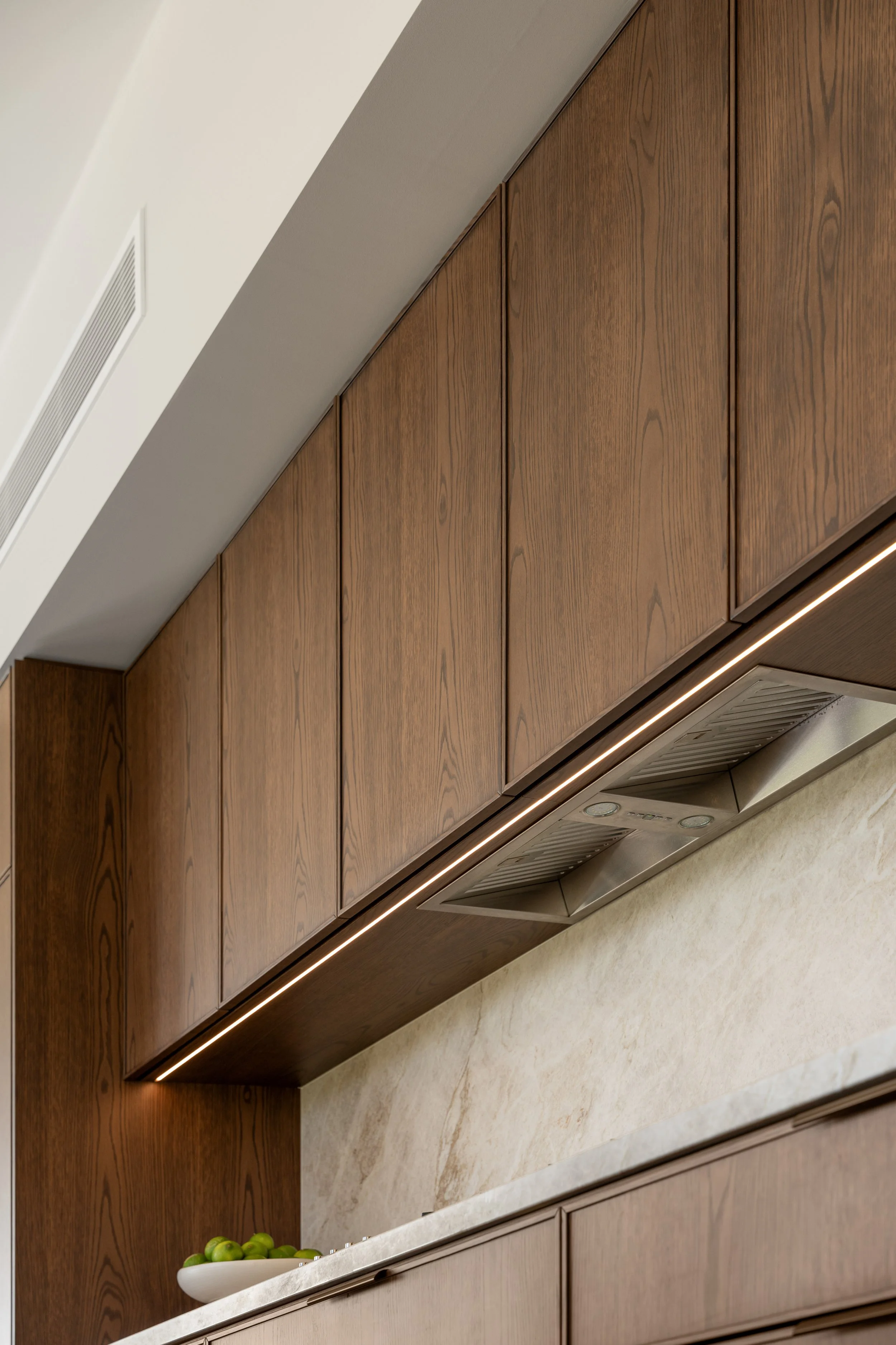 View of kitchen cabinetry with a marble countertop, wooden upper cabinets, a built-in range hood, and a fruit bowl with green apples.