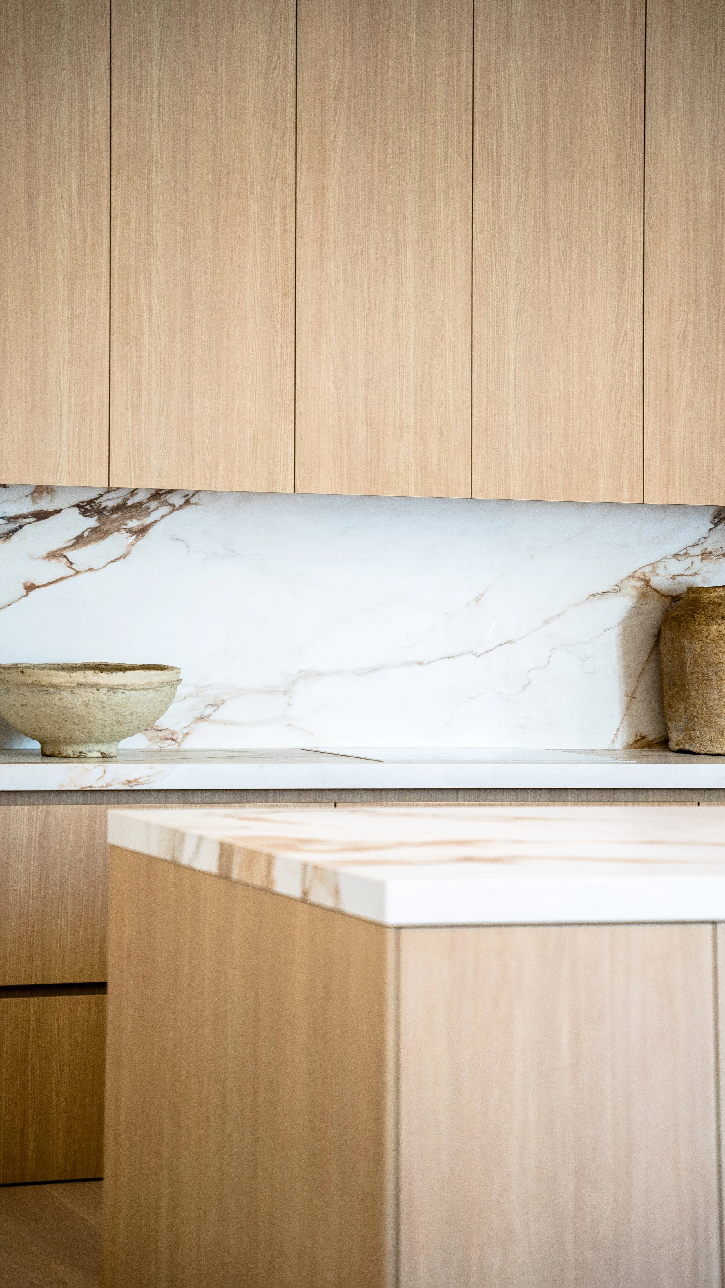 Modern kitchen with light wood cabinets, white marble backsplash with brown veining, and a marble countertop with wooden accents, decorated with two ceramic bowls and a rustic clay pot.