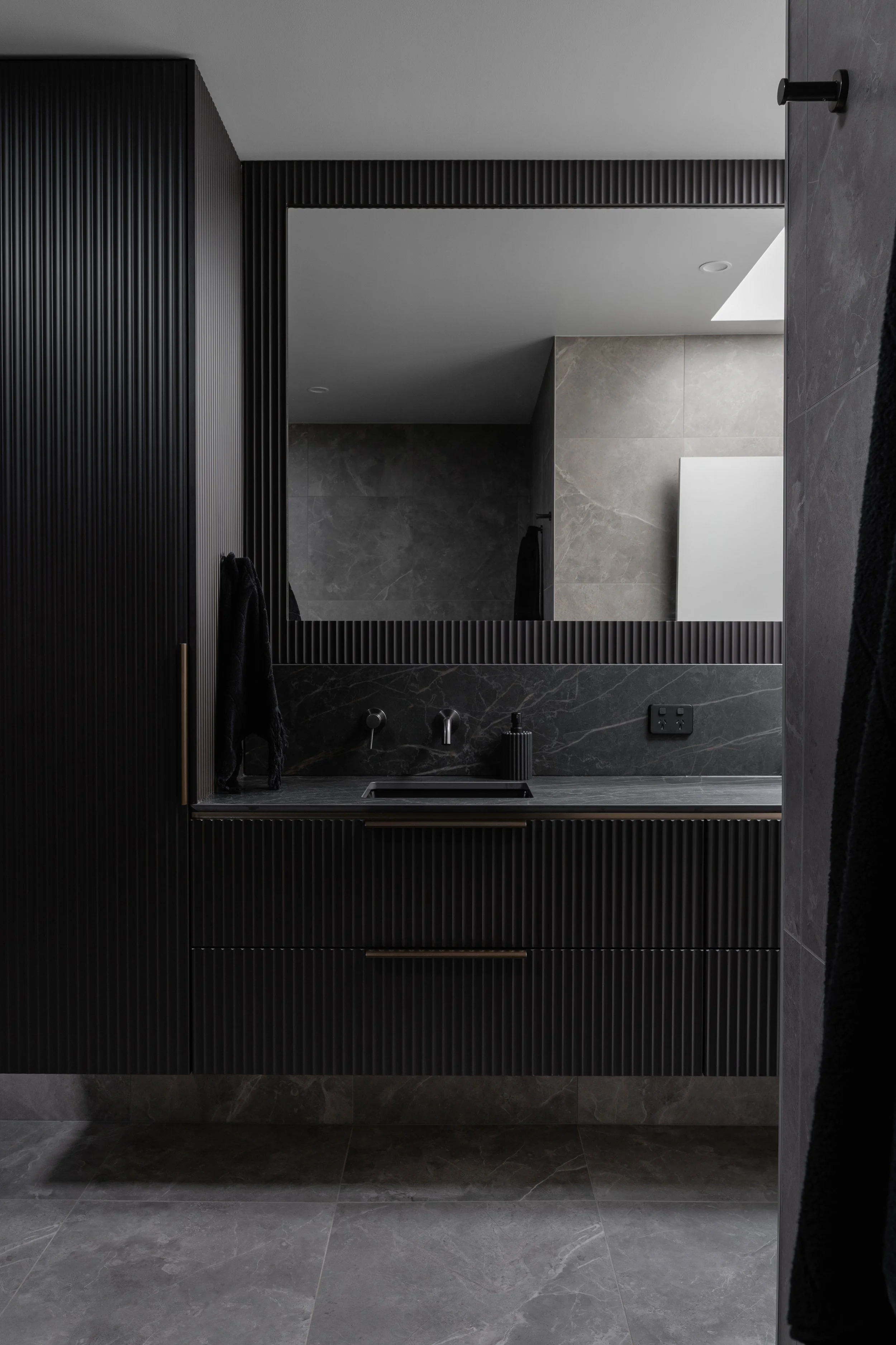 A modern bathroom featuring a large mirror with a black ribbed frame and a black sink and faucet. The walls and floor are tiled in dark gray marble, and there are black towels hanging on the wall. The ceiling has recessed lighting and a skylight lett
