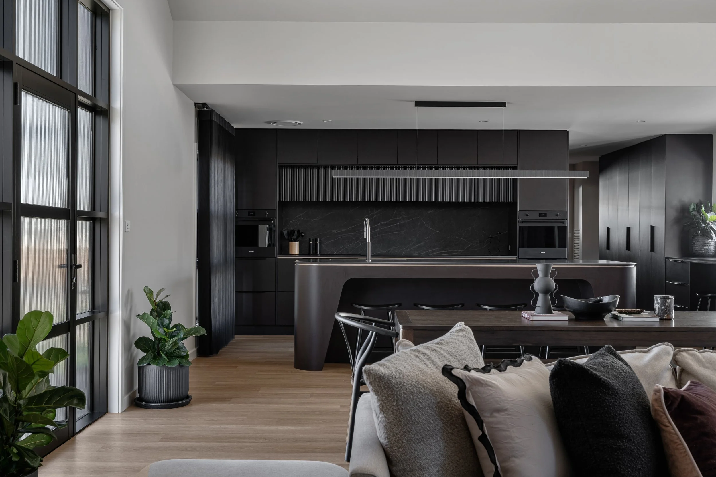 Modern kitchen with black cabinetry, a black marble backsplash, and a kitchen island with a metallic faucet. Nearby, a dining table is partially visible, with several chairs and decorative items. Large windows with black frames allow natural light, a