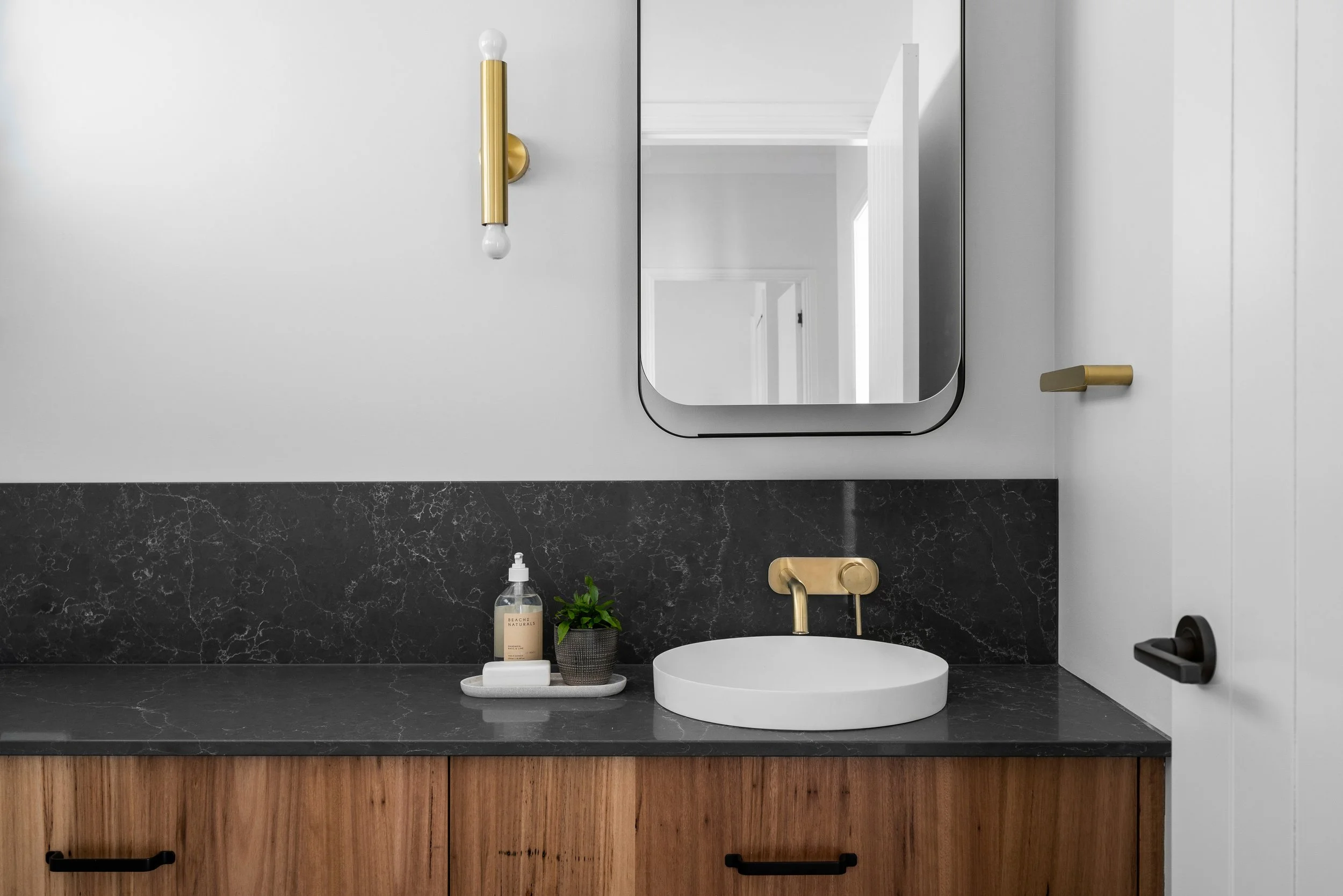 Modern bathroom with a black marble countertop, round white sink, gold faucet, mirror, wall sconce, and wooden cabinet below.