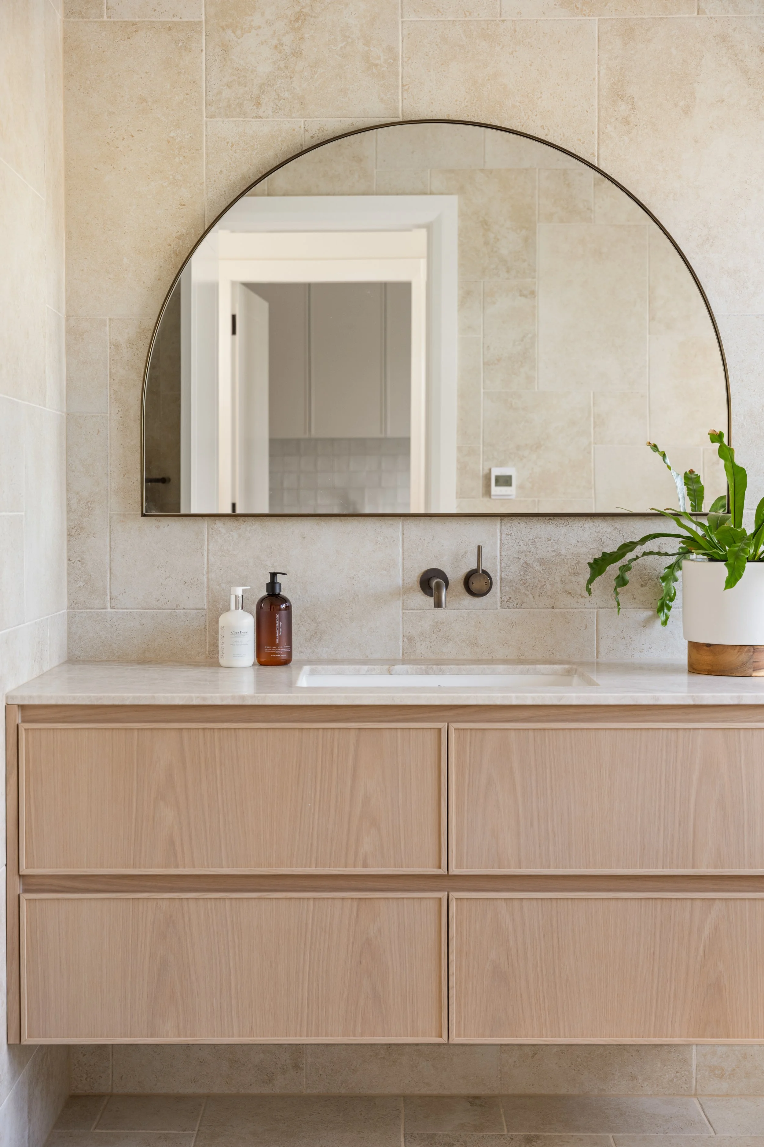 Bathroom vanity with a mirror, countertop, and bottles of soap or lotion, with beige tiled walls and a potted plant on the right.