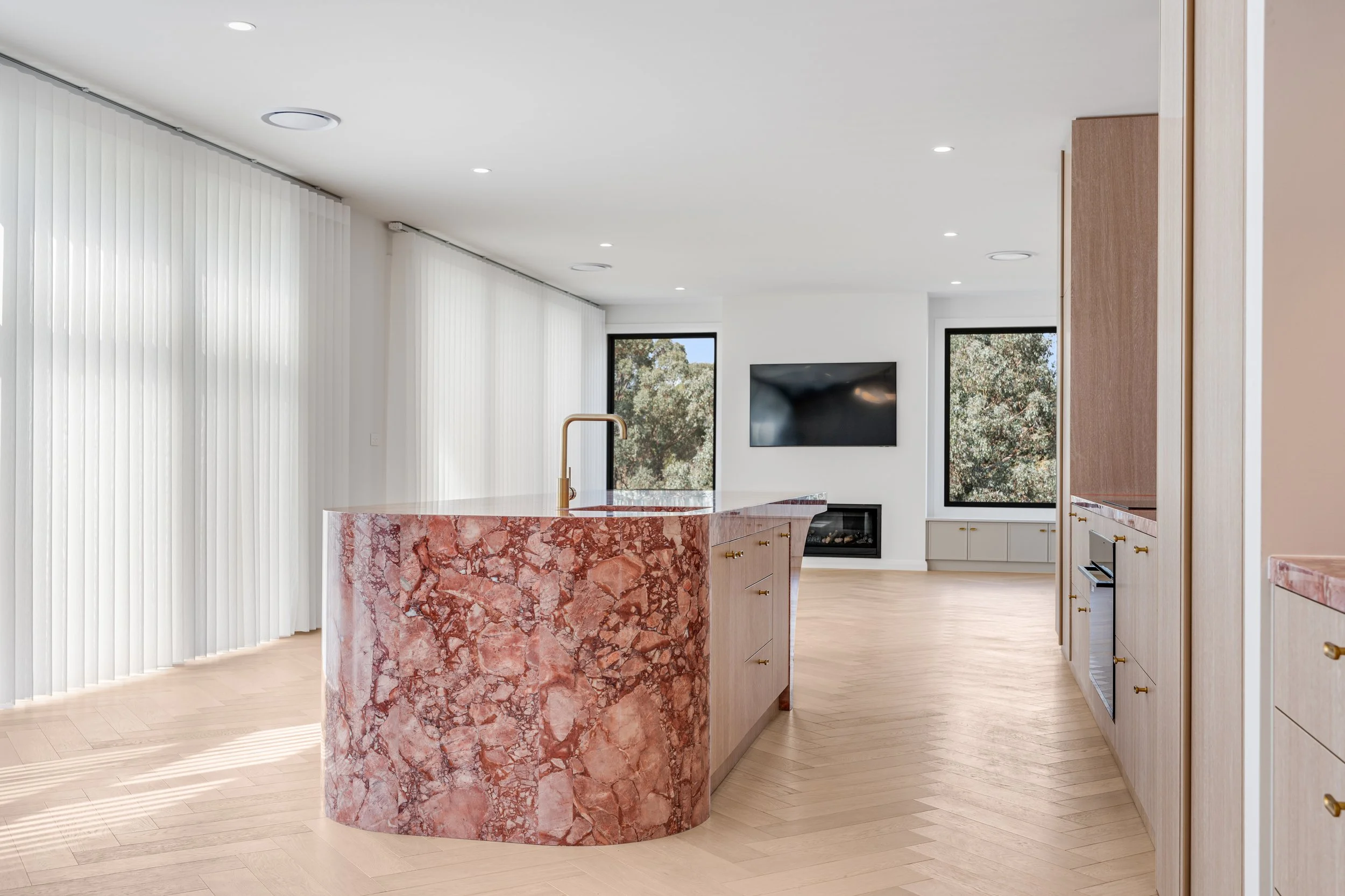 Modern kitchen with pink marble island, white cabinets, built-in fireplace, flat-screen TV, large windows, white curtains, light wood flooring, and minimal decor.