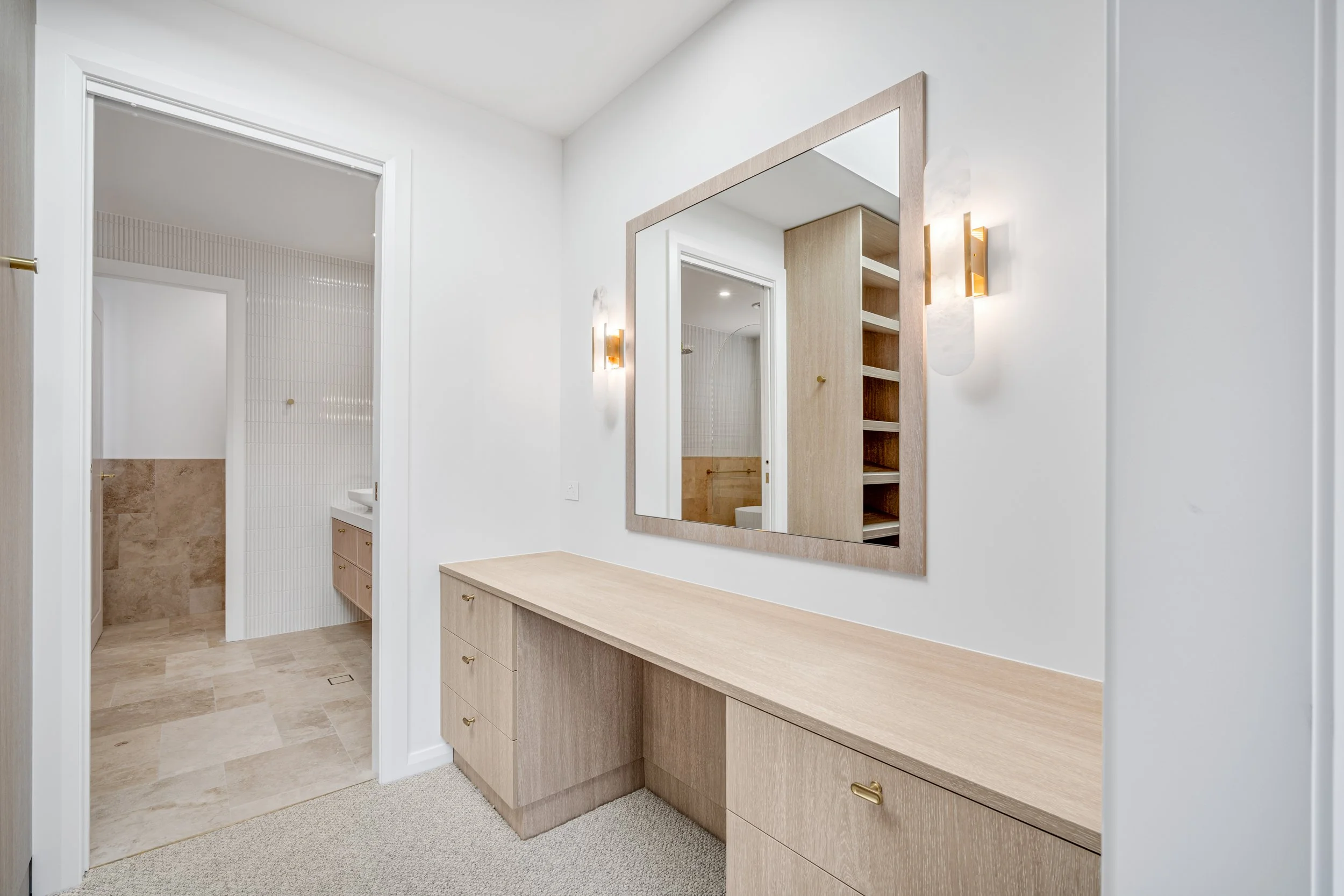 Interior of a modern bedroom with a large mirror, wooden dresser, and wall-mounted light fixtures. A doorway leads to a bathroom with beige tiles and a shower.