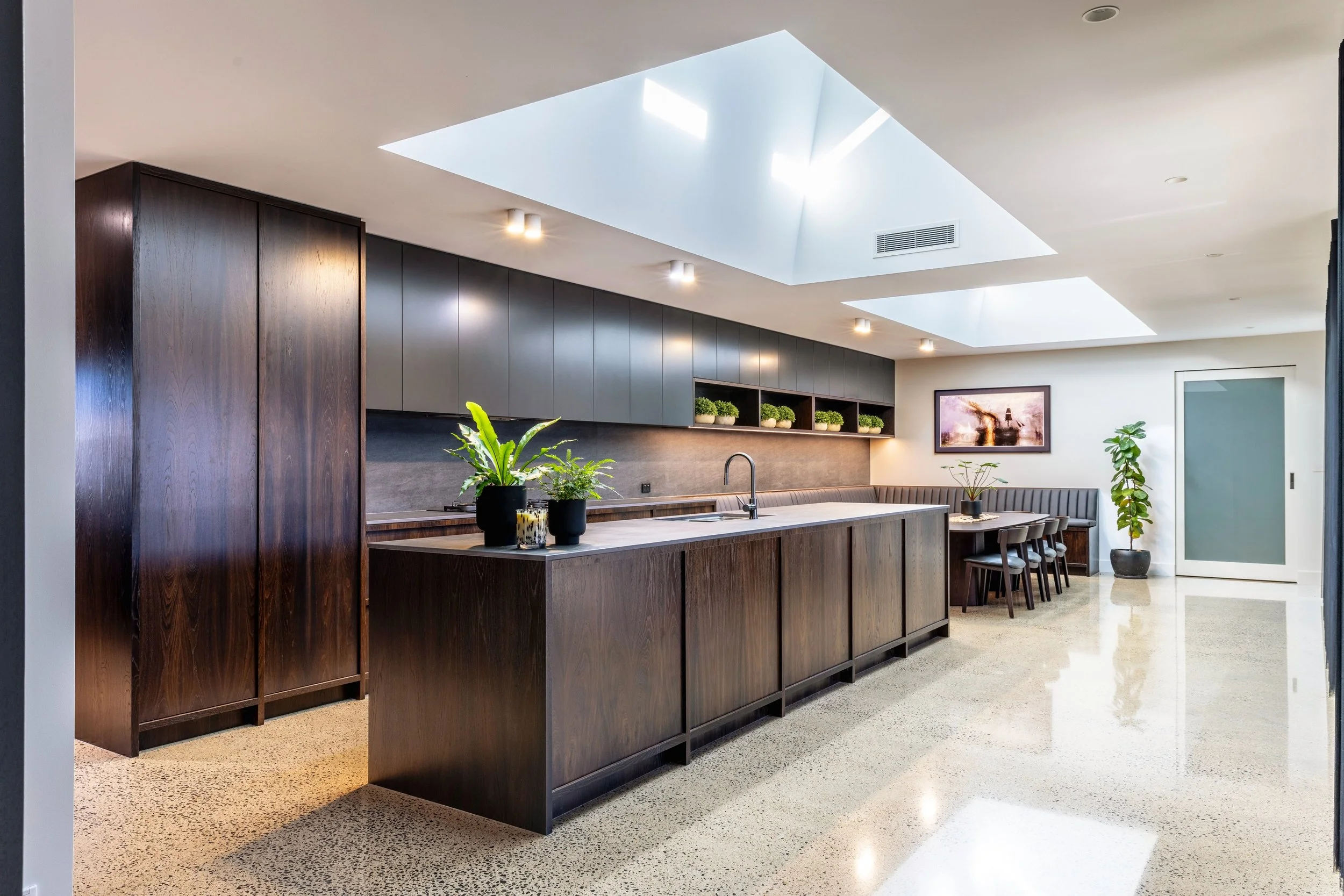 Modern kitchen with skylights, dark wood cabinets, a long island with plants, and a dining area with chairs and artwork.