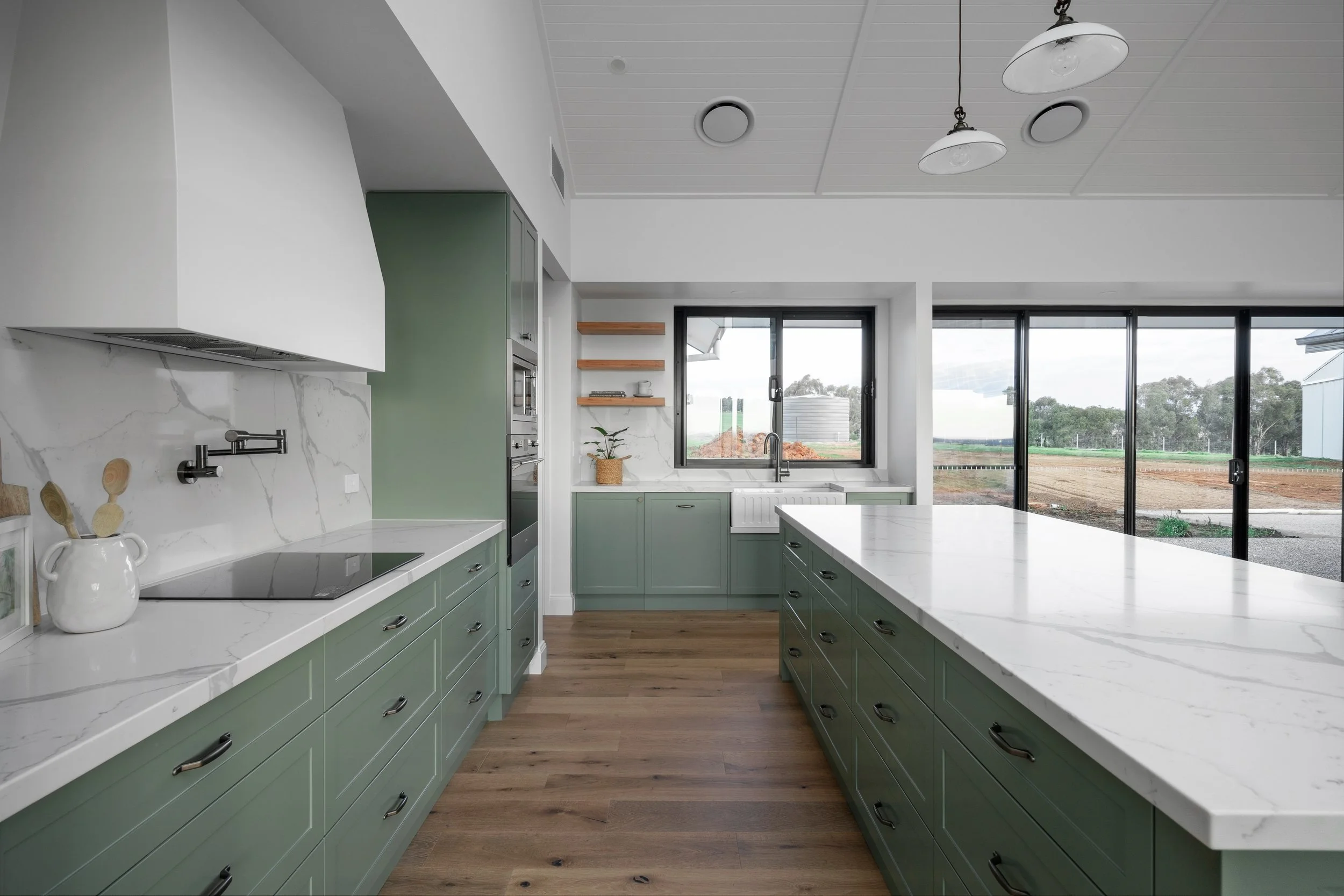 Modern kitchen with green cabinets, white marble countertops, and large windows overlooking a rural landscape with trees and fields.