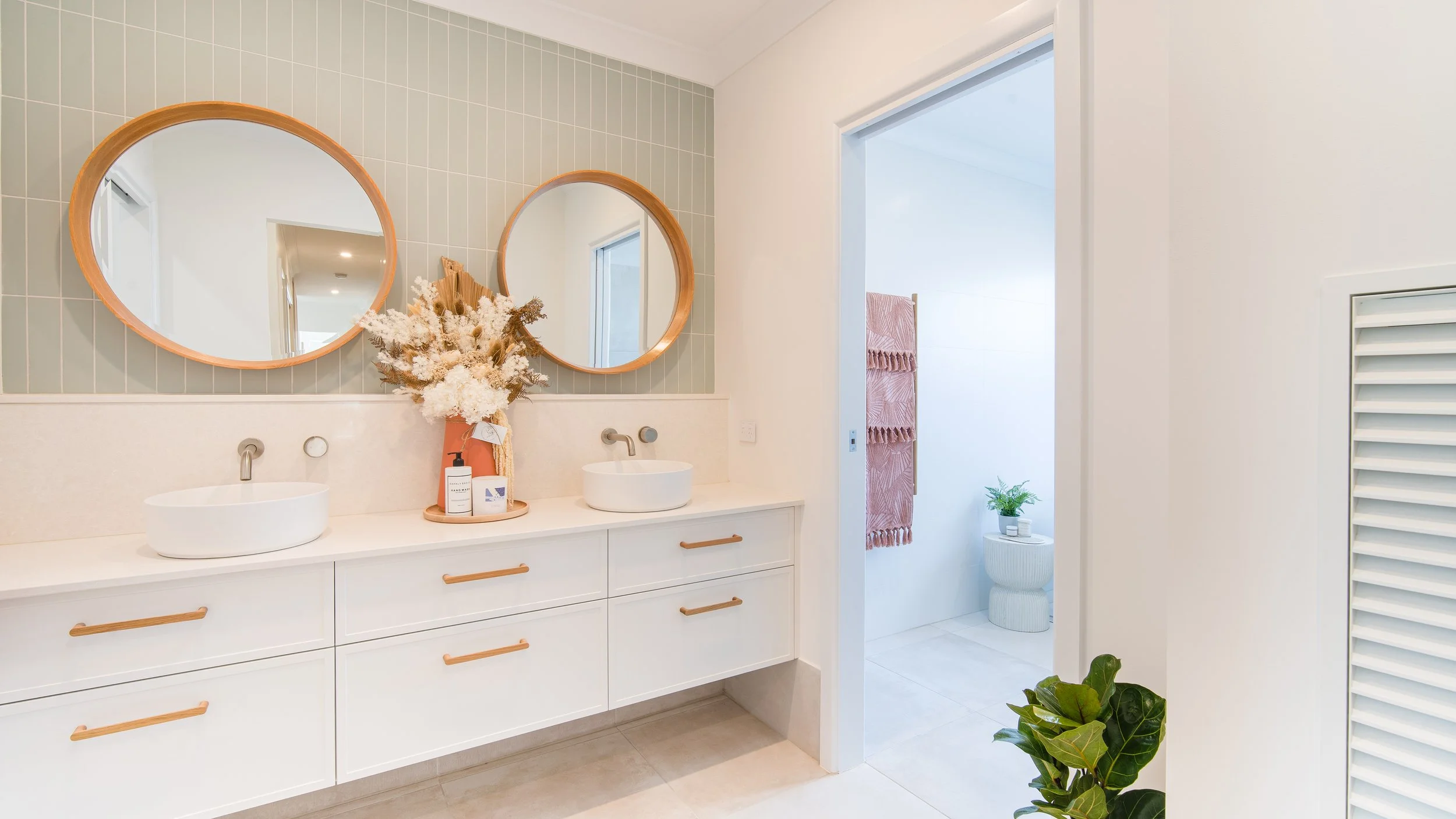 Modern bathroom with a white double vanity featuring round vessel sinks, wooden handles, and a vase with dried flowers. Two round mirrors hang above the sinks. In the background, a separate toilet area with a pink towel and a small table with a green