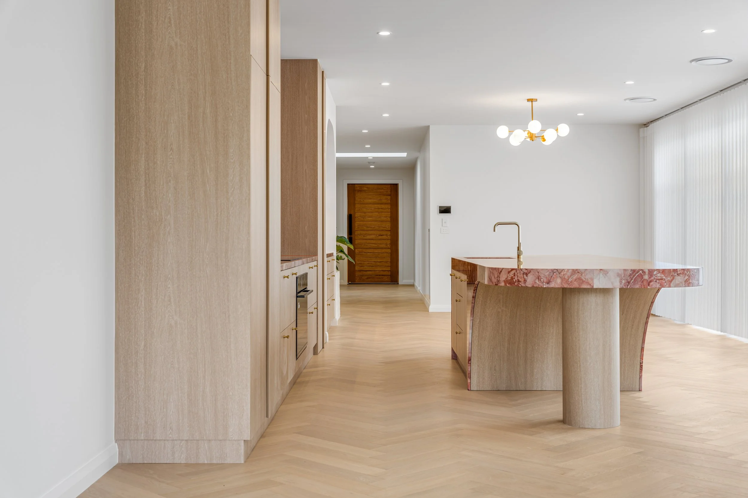 Modern interior space with light wooden flooring, white walls, and minimalist decor. A room features built-in wooden cabinets, a wooden door, a pink marbled kitchen island with a gold faucet, a contemporary chandelier, and vertical curtains covering 