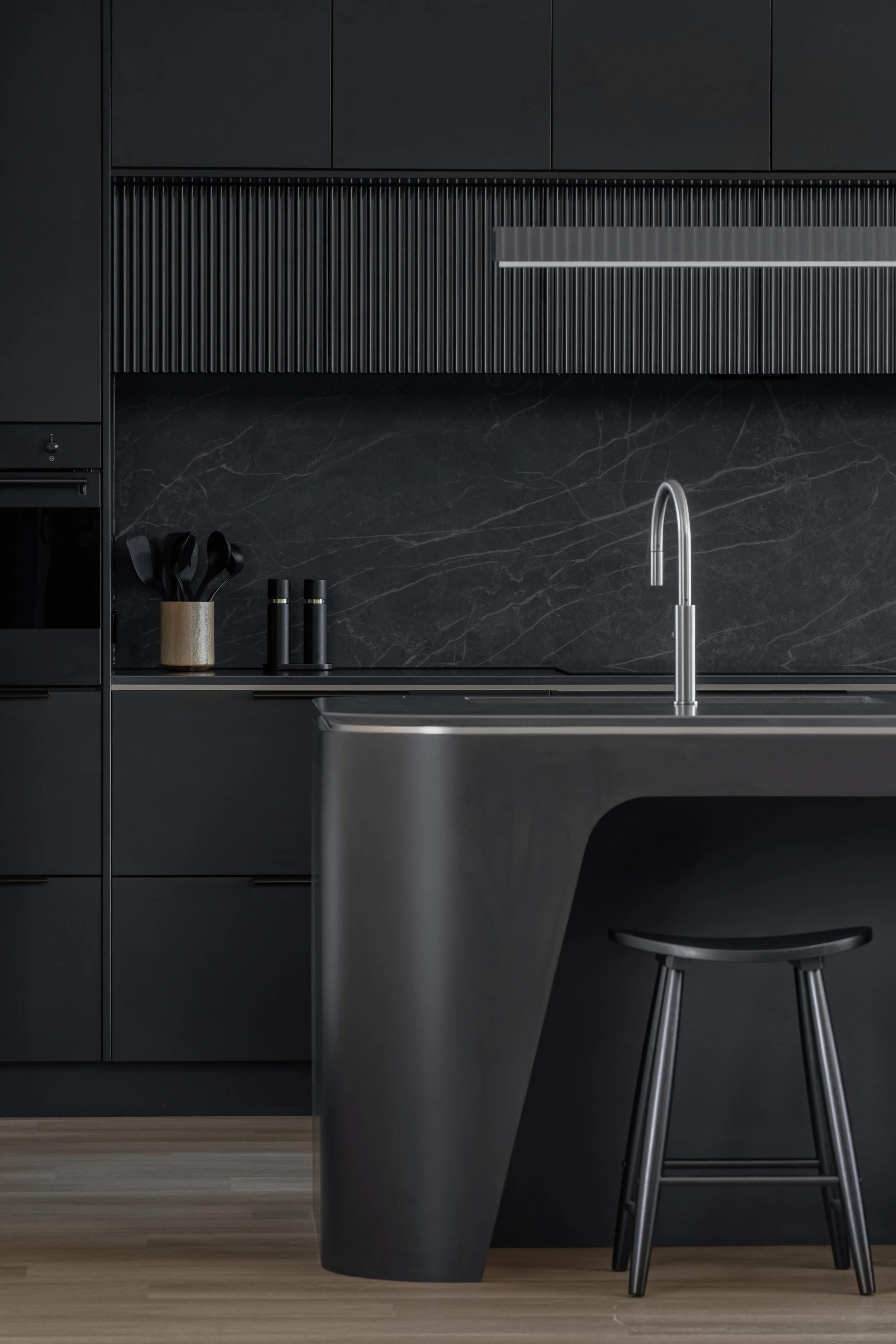 Modern kitchen with black cabinetry, black marble backsplash, stainless steel faucet, black utensils in a wooden holder, and a black barstool.