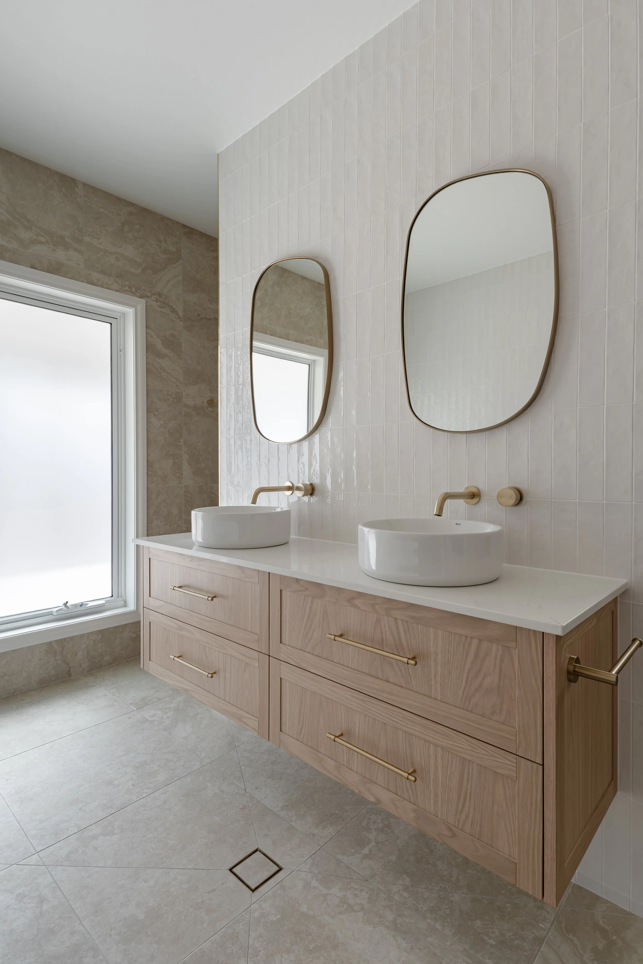Modern bathroom with a light wood vanity, double vessel sinks, oval mirrors, a large window, and beige tiled walls and floor.