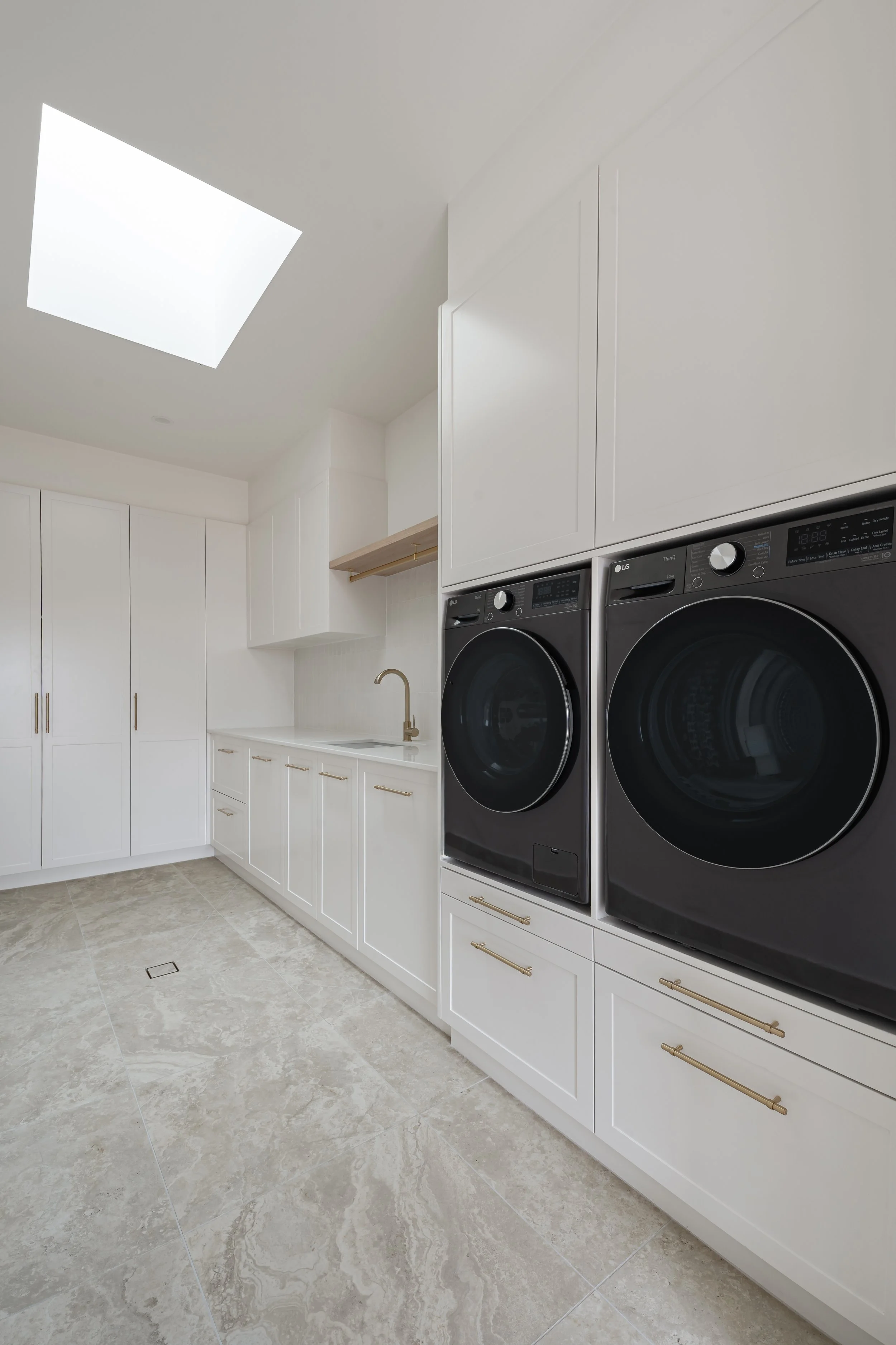 Modern laundry room with white cabinetry, a built-in sink, and black front-loading LG washer and dryer set, beige marble flooring, and a ceiling skylight.