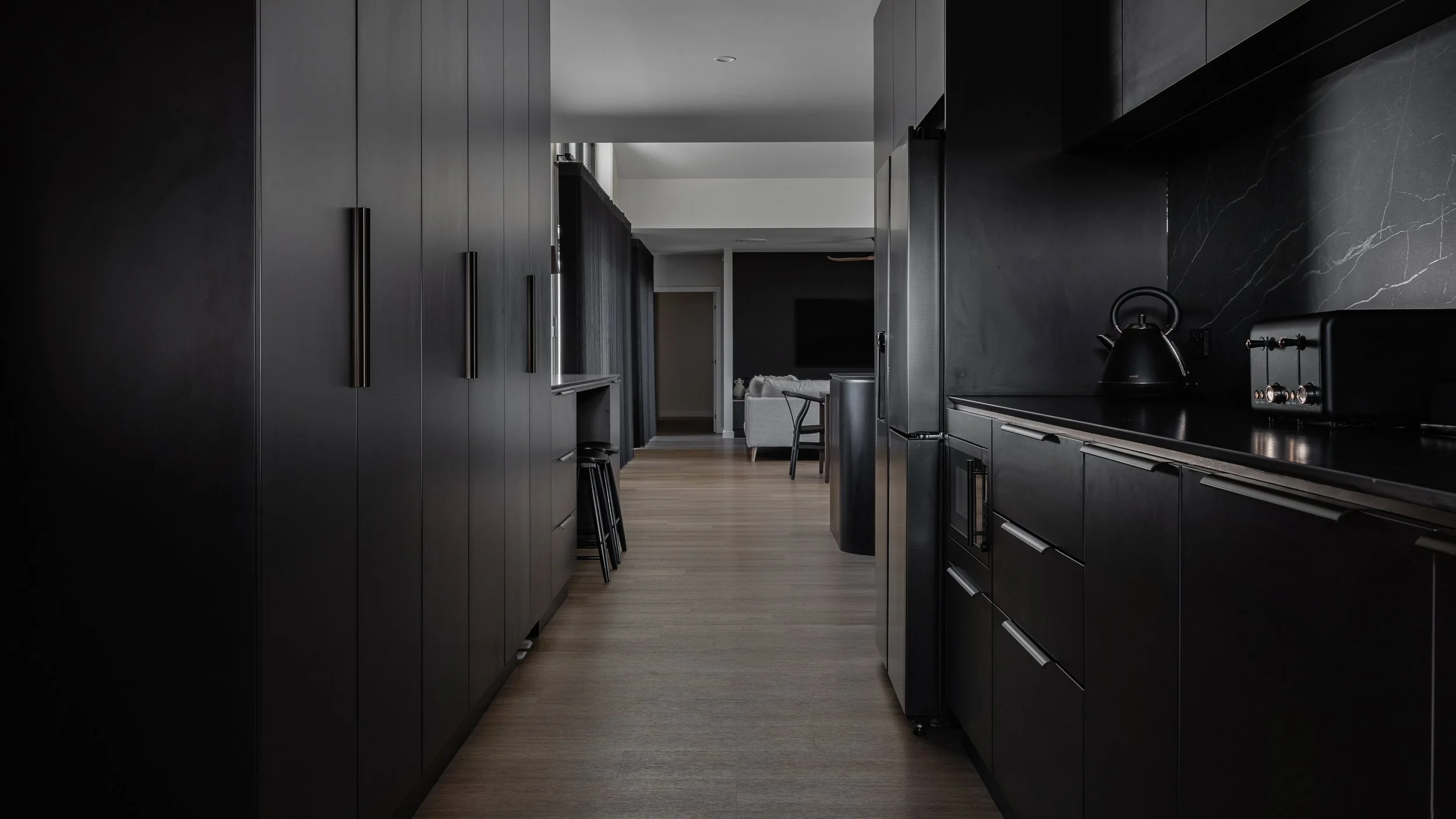 A modern kitchen with black cabinetry and a black countertop, featuring a kettle and toaster, with a view into a living area with a sofa in the background.