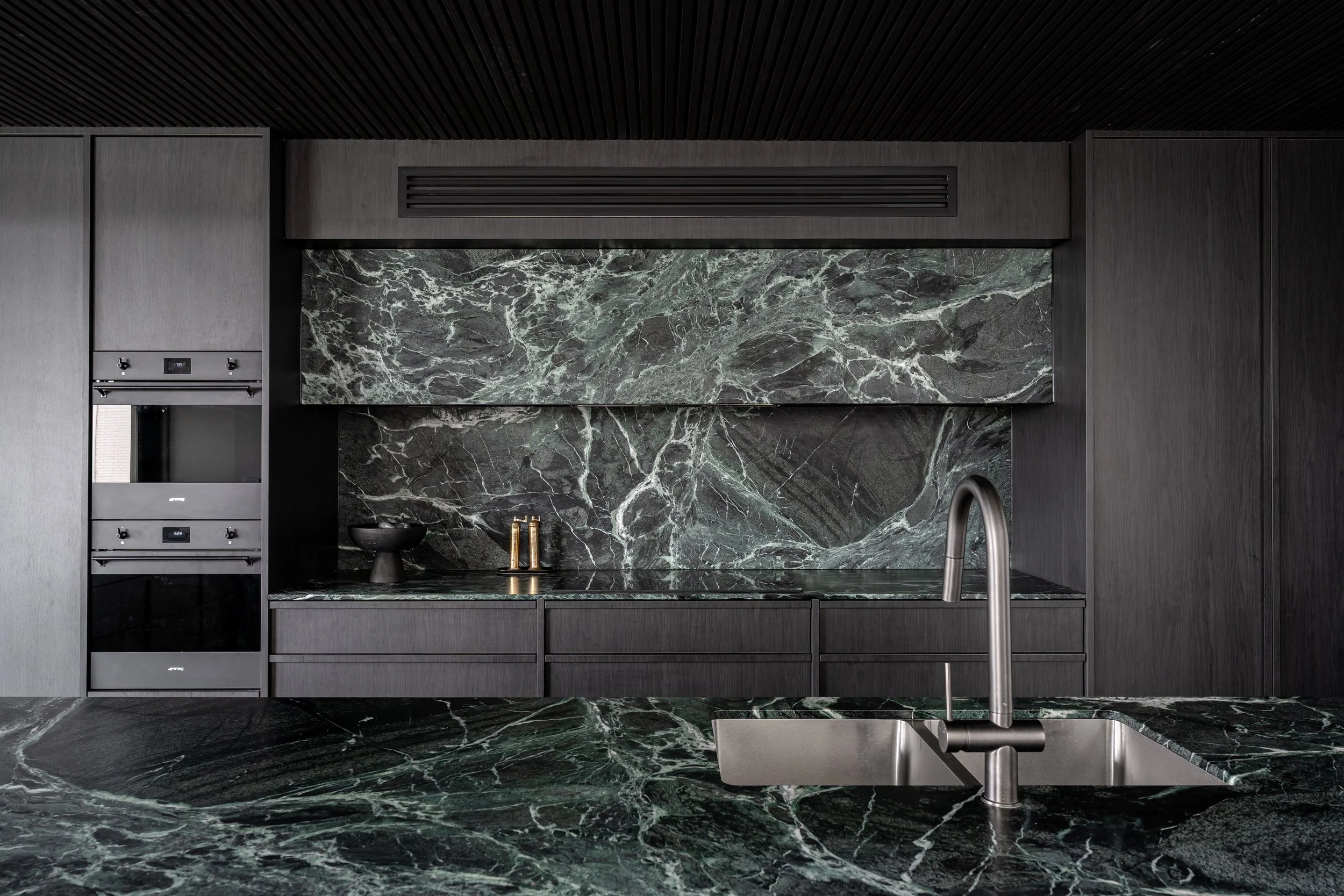 Modern kitchen with dark gray cabinets and walls, black marble backsplash and countertop with white veining, stainless steel sink and faucet, built-in oven and microwave, and ceiling with black slats.