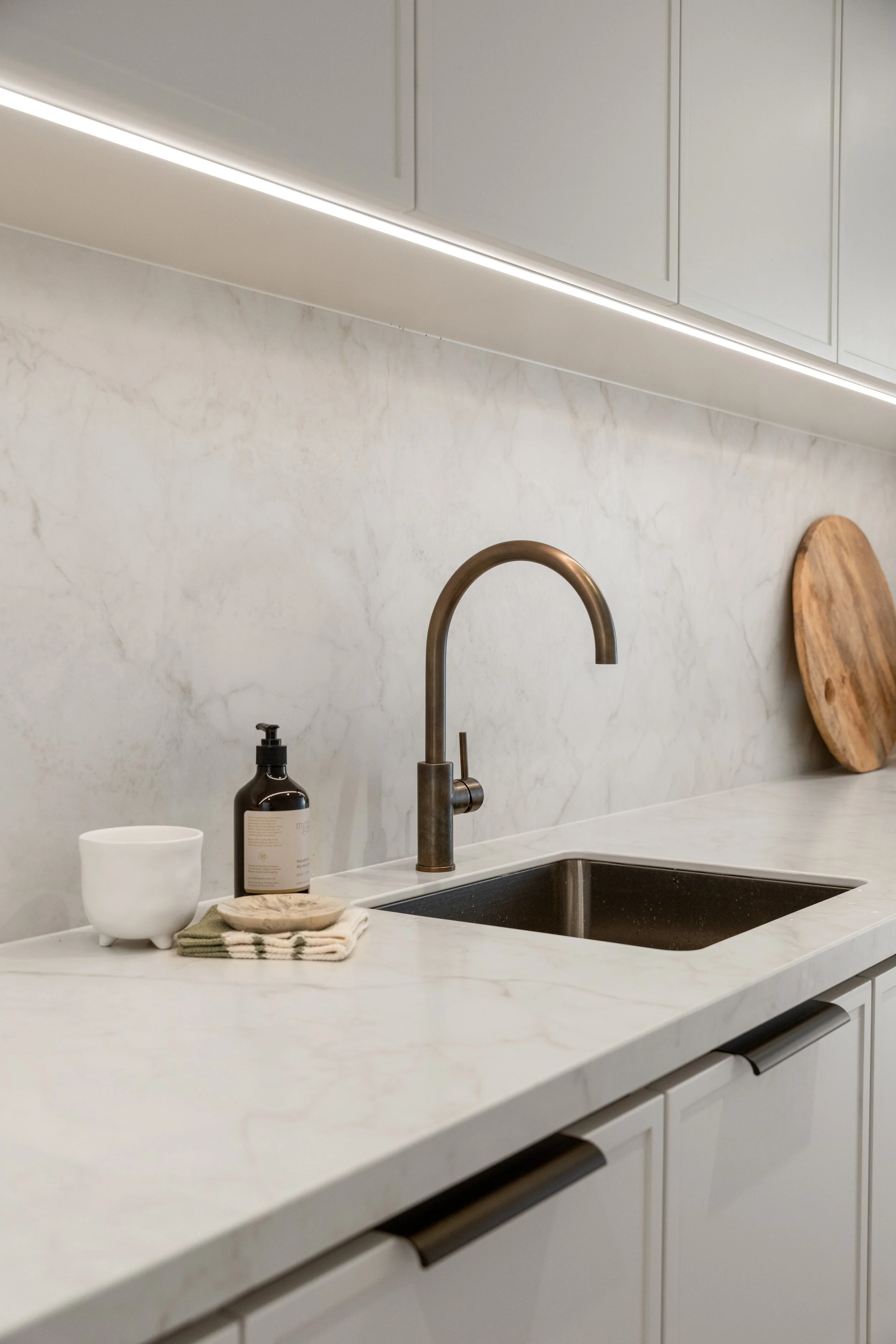 A modern kitchen countertop with a white marble surface, a stainless steel sink, a brushed bronze faucet, and a white backsplash. There are decorative items including a white bowl, a brown soap dispenser, and a wooden cutting board leaning against the backsplash.