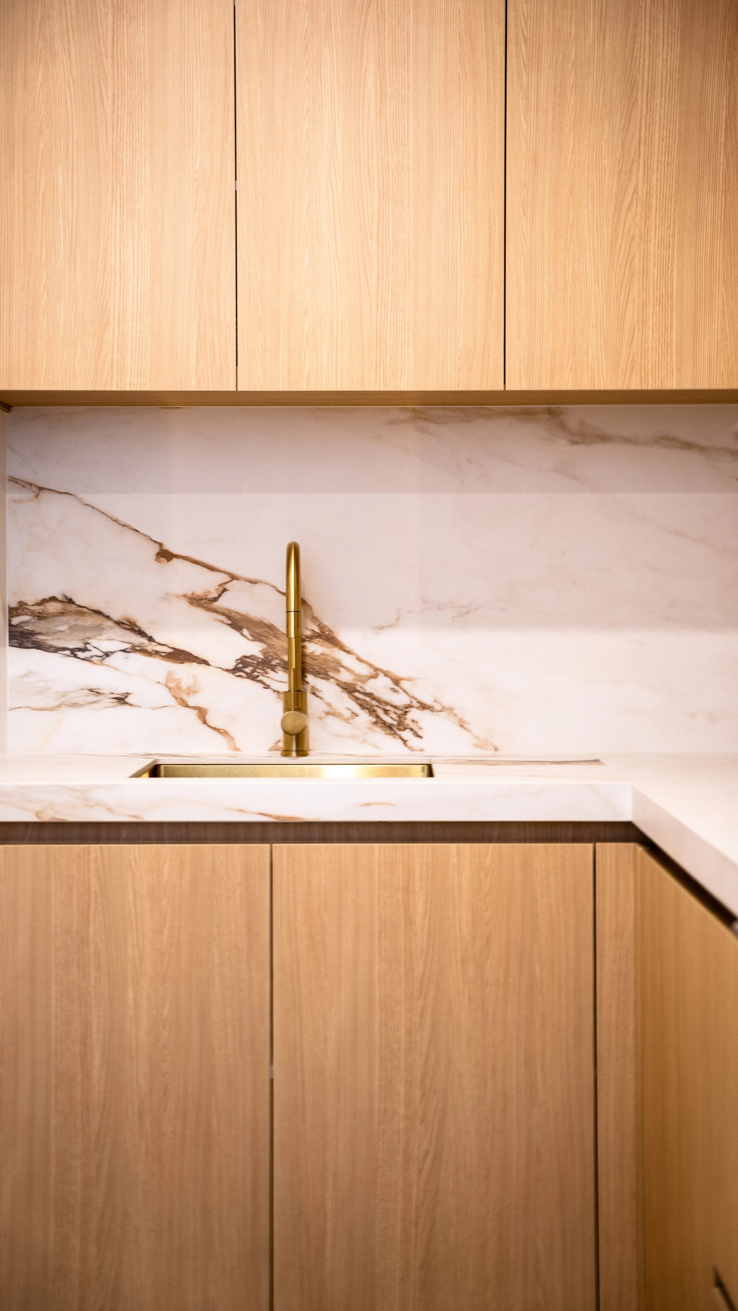 Kitchen with light wooden cabinets, marble countertop and backsplash, and a gold-colored faucet.
