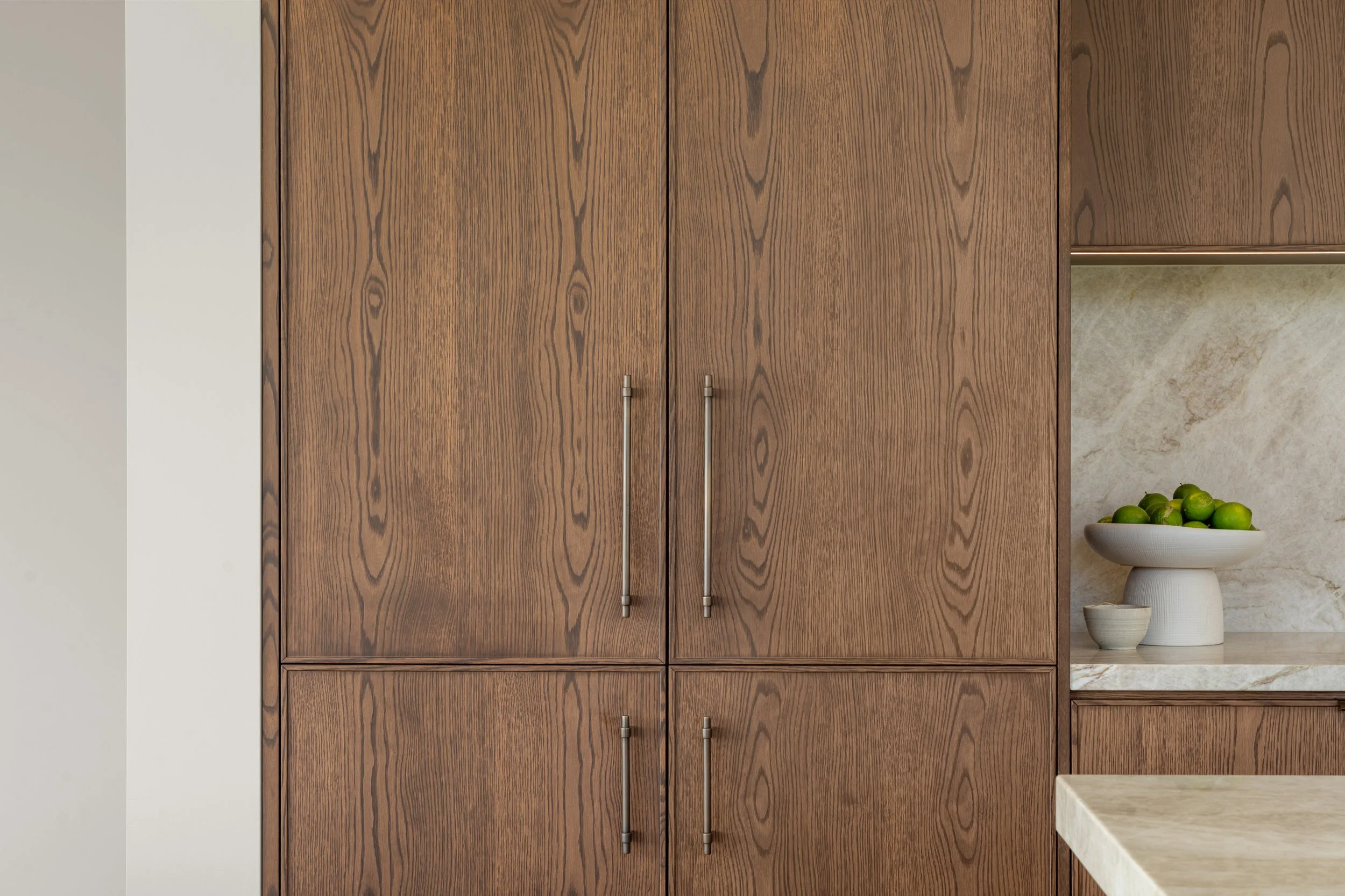 Wooden cabinets with metal handles and a glimpse of a beige marble countertop with a bowl of green limes.