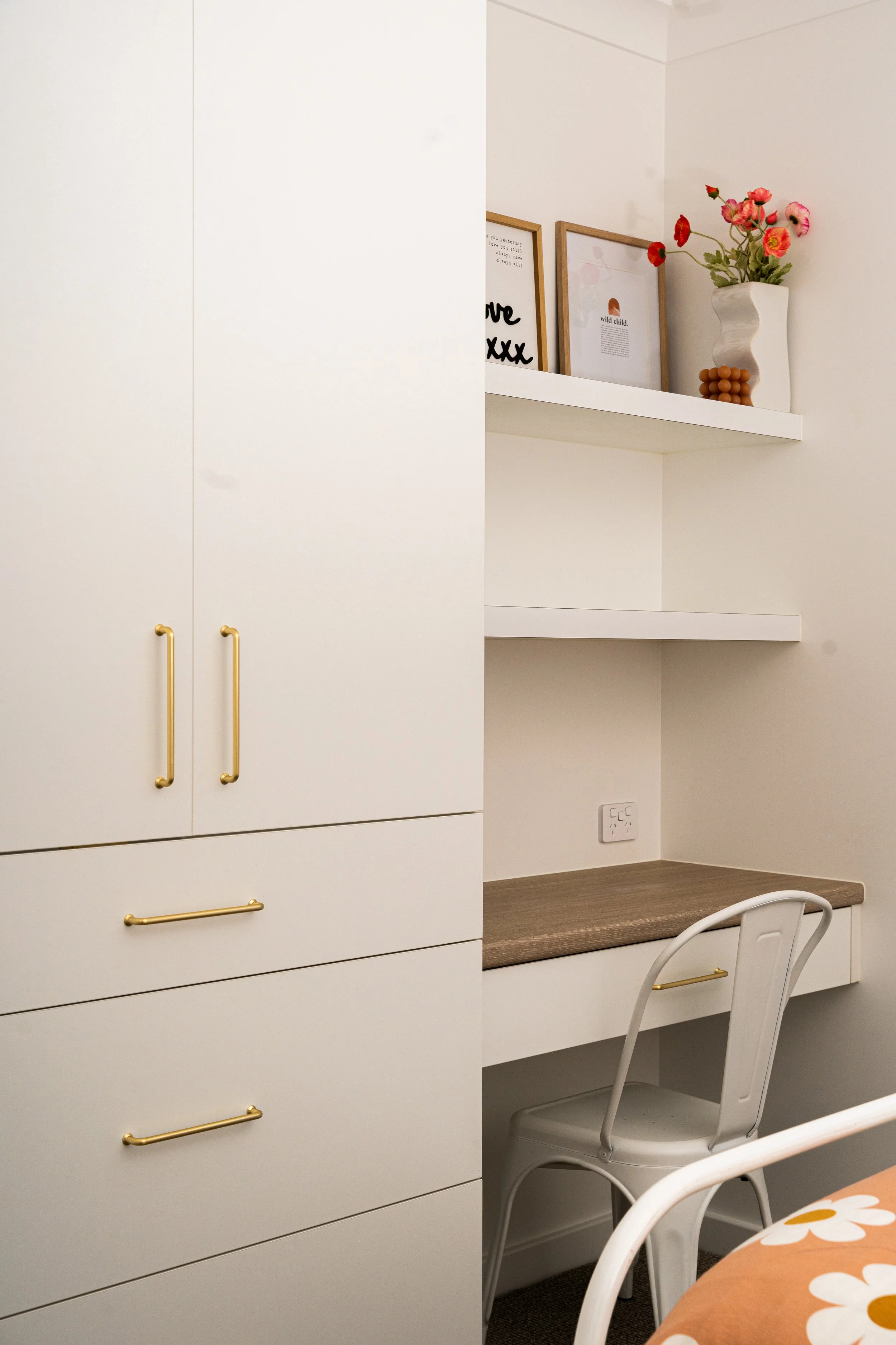A section of a room with white cabinetry featuring gold handles, a built-in desk with a brown surface, white chair, and wall shelves holding framed art, decorative objects, and a tall white vase with pink and red flowers.