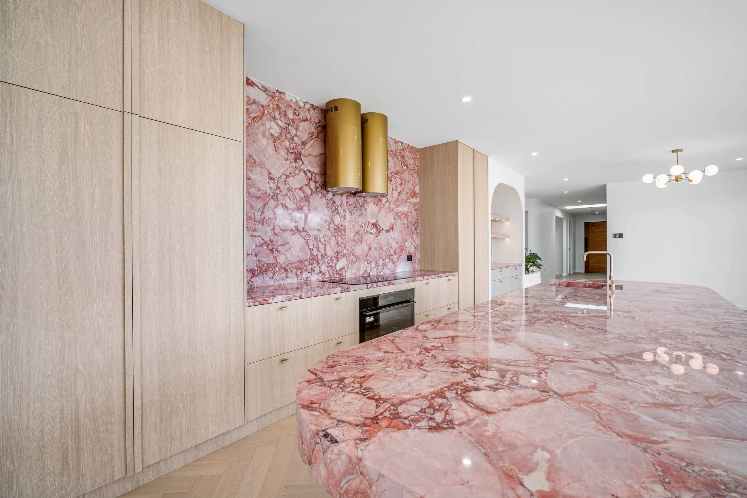 Modern kitchen with pink marble countertops, wooden cabinetry, and a pink marble backsplash. Gold cylindrical range hoods hang above the stove, and a pendant light fixture is visible in the background.