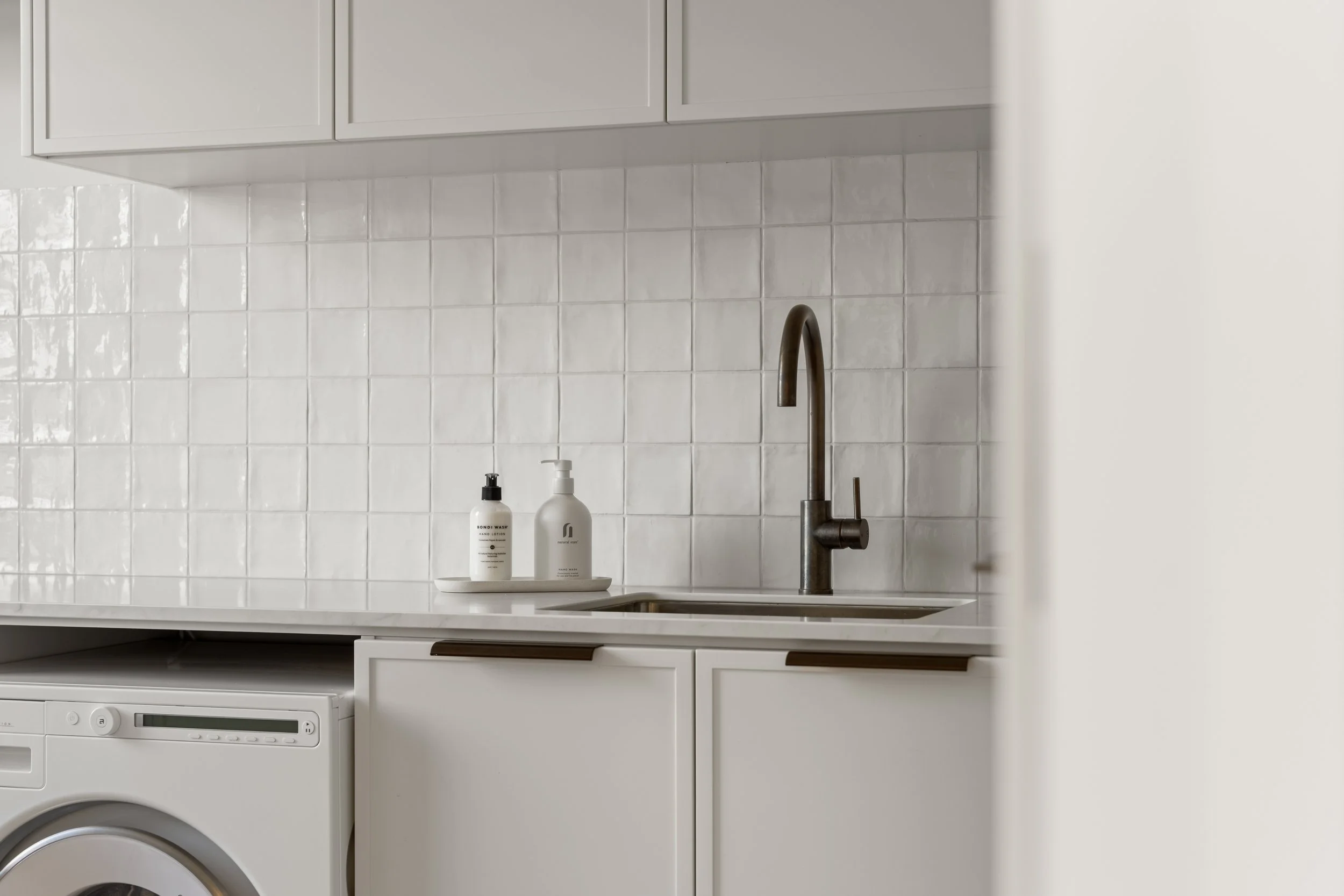 A white kitchen with white cabinets, a silver faucet at a sink, backsplash tiled in white, and two soap dispensers on a small tray by the sink, with a washing machine below.