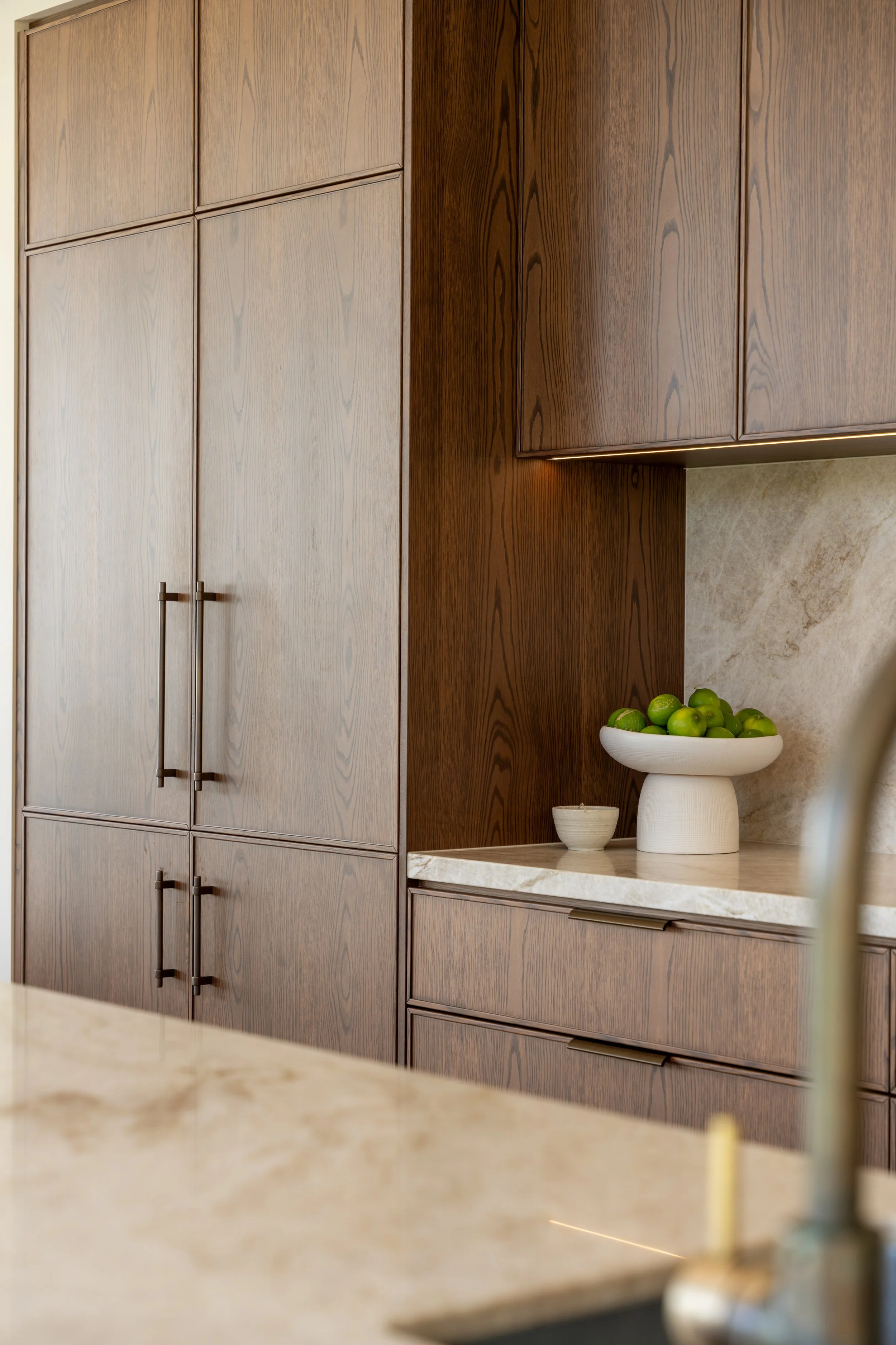 Wooden kitchen cabinets with black handles, beige marble countertop, white bowl of green limes, and a stone backsplash.