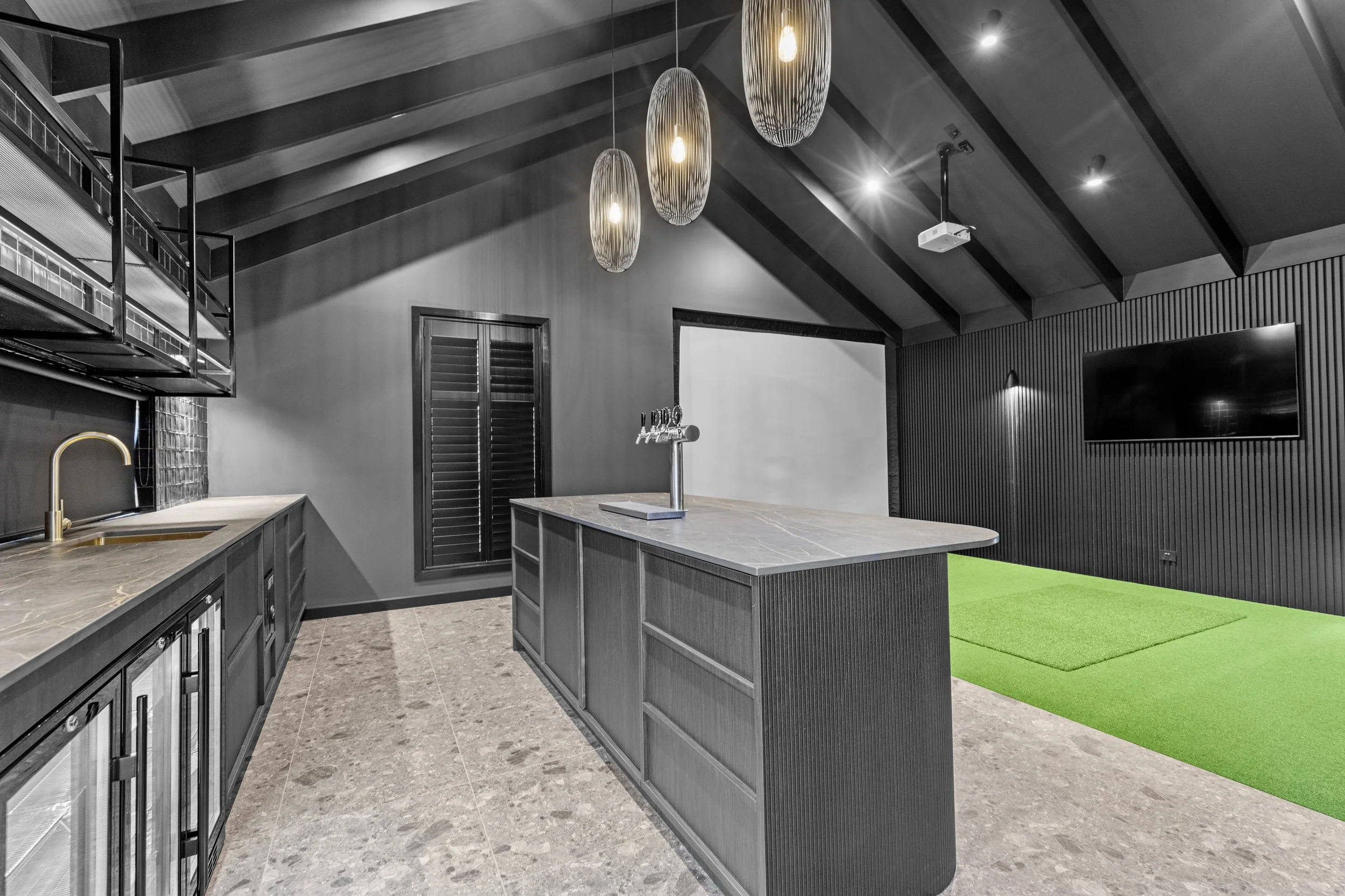Modern kitchen with black cabinetry, marble countertops, and hanging pendant lights. There is a green carpeted area and a black slatted wall with a mounted flat-screen TV.
