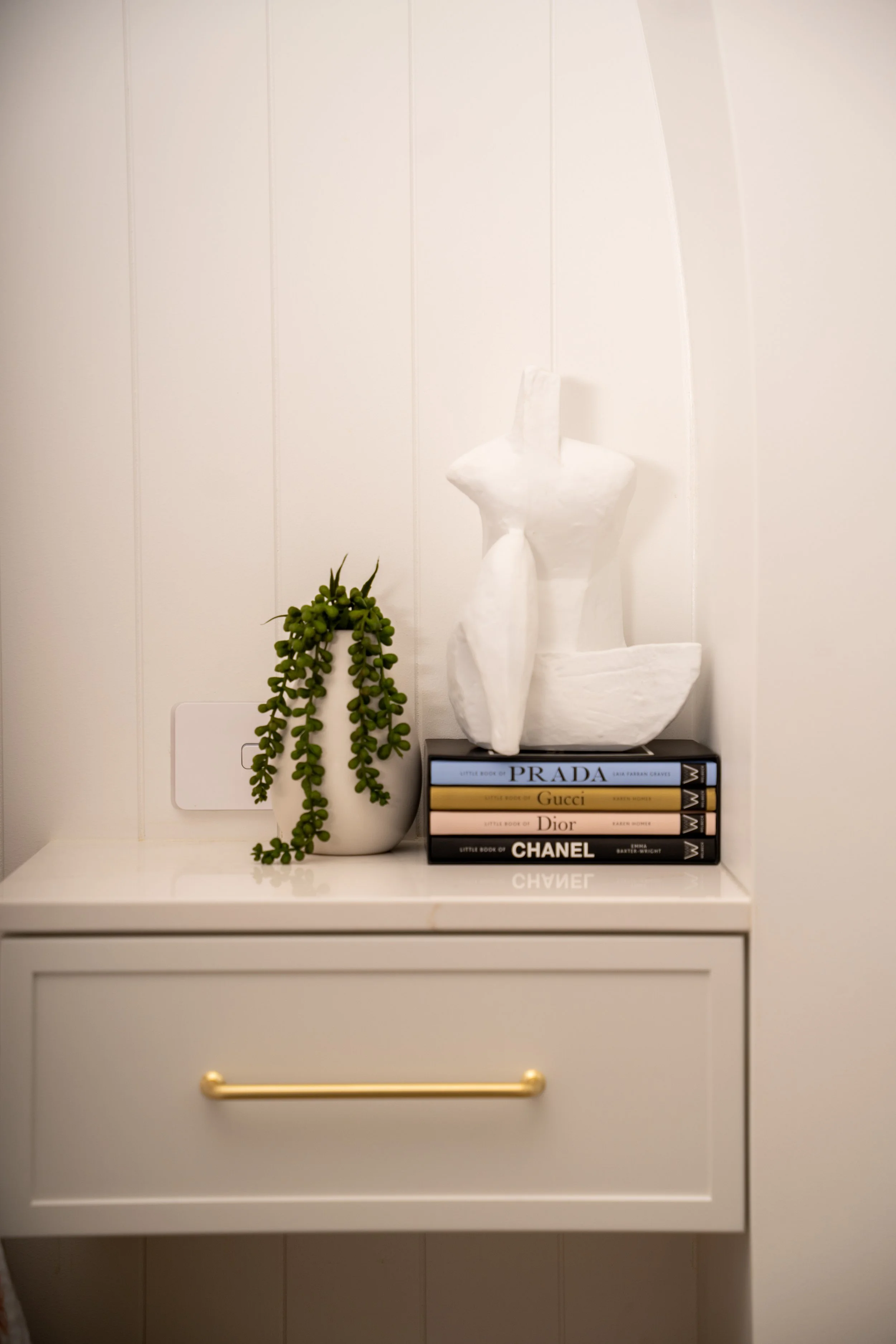 Decorative white sculpture resembling a stylized bird placed on top of a stack of fashion books, next to a small green trailing plant in a white pot on a white cabinet.