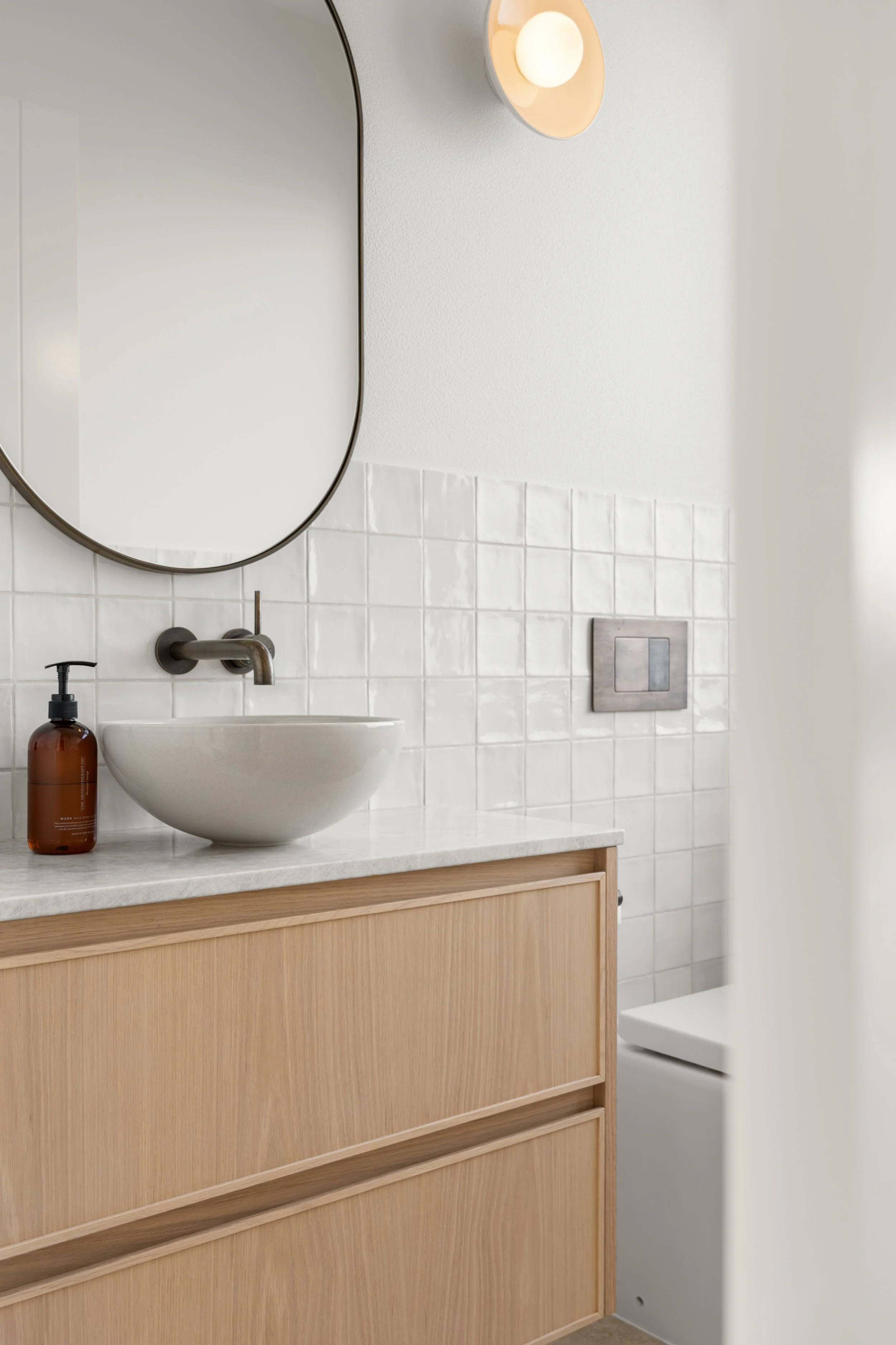 A modern bathroom vanity with a round vessel sink, a wall-mounted faucet, a brown soap dispenser, and a large oval mirror. The vanity has a light wood finish, and the wall behind features white tiles and a textured white wall with a light fixture.