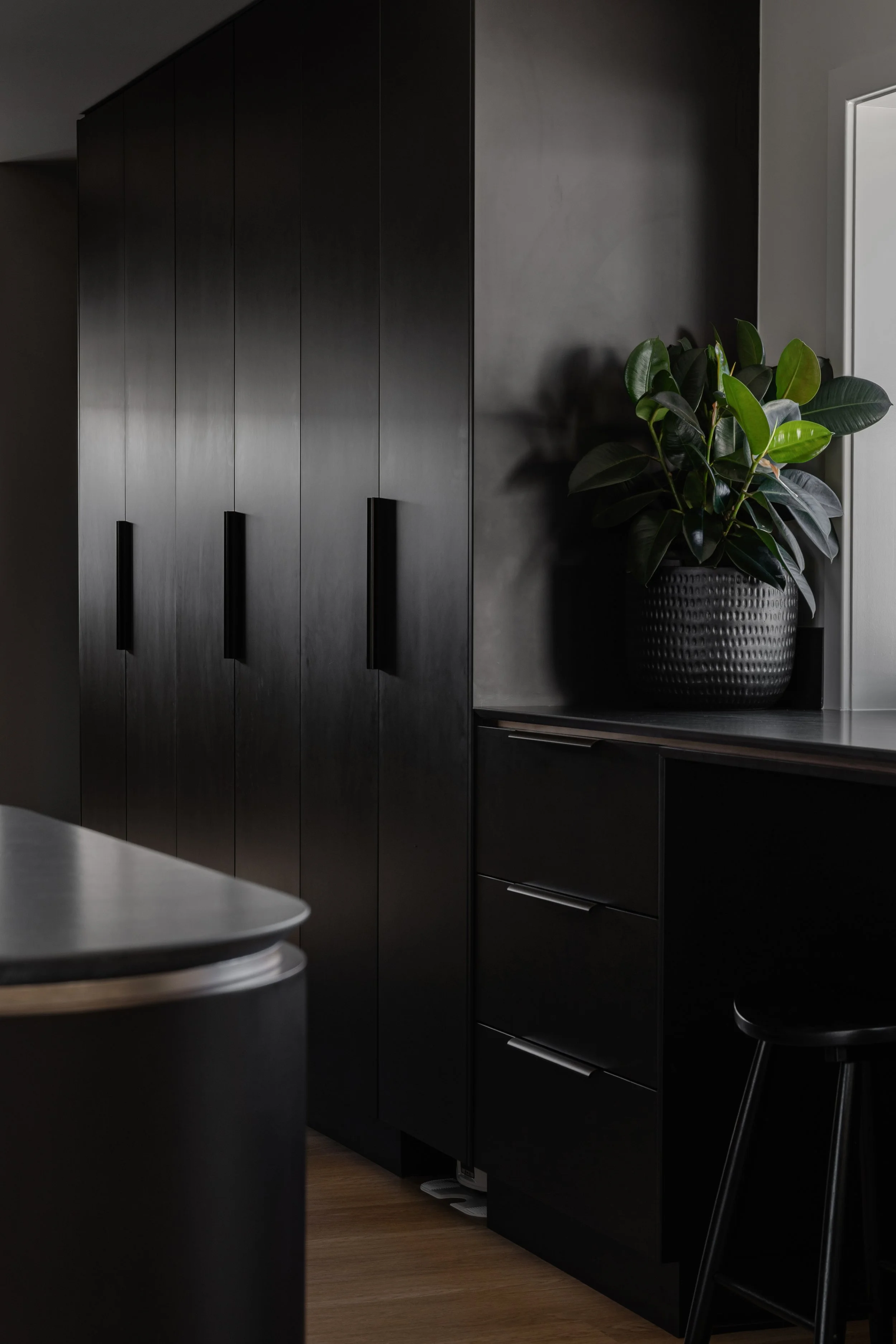 A modern black cabinet with three handles, a potted green plant on top, and a partial view of a gray chair in a room with wooden flooring and light gray walls.